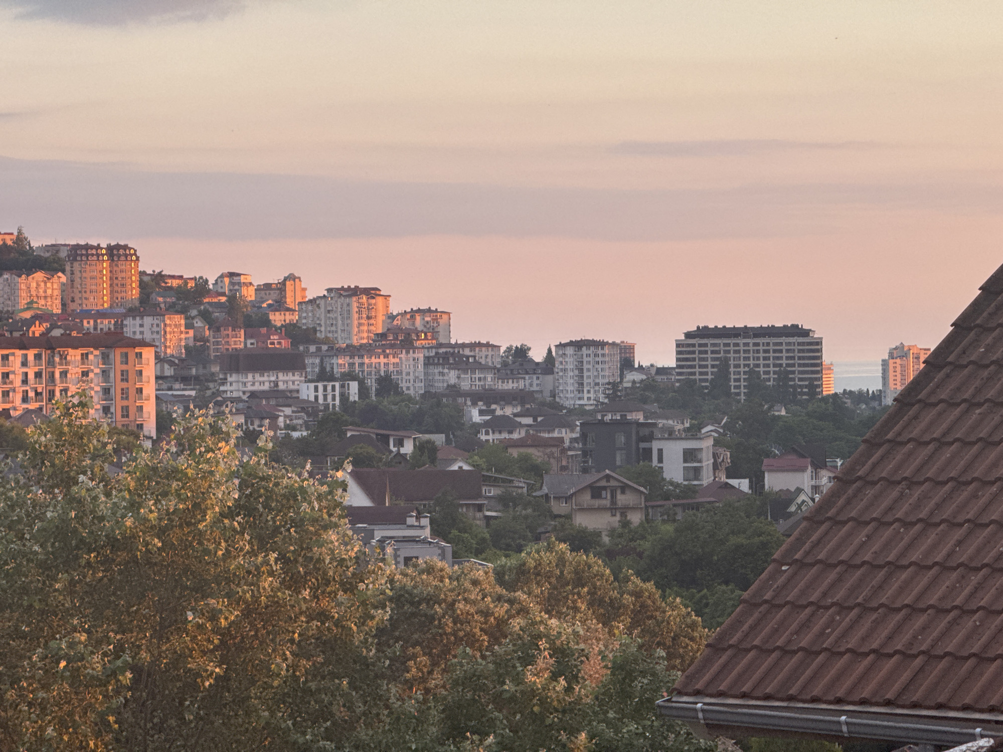A panoramic view of a coastal city, likely Sochi, Russia, is captured during the golden hour of sunset. The sky is awash with soft hues of pink and orange, indicating the sun is low on the horizon. A dense urban landscape unfolds across rolling hills, characterized by a mix of apartment buildings, houses, and trees.
In the foreground, the terracotta tiled roof of a building dominates the lower right corner, suggesting the viewpoint is from an elevated position. Lush green trees with sunlit foliage spread across the mid-ground, with some trees showing warmer, autumnal colors, possibly due to the light or seasonal change.
The cityscape is dotted with various architectural styles. Several multi-story apartment buildings, some with balconies and a grid-like facade, are prominent. One particularly tall, modern building with a dark, flat roof stands out in the mid-right section of the image. Smaller, more traditional houses with pitched roofs are nestled among the larger structures and trees. The presence of what appears to be a body of water or the sea in the distance, visible between buildings on the right side, further reinforces the coastal context.
The scene is tranquil, bathed in the warm, diffused light of sunset. The overall mood is peaceful and serene, with the cityscape appearing calm and settled as the day draws to a close. There is no visible text in the image.
