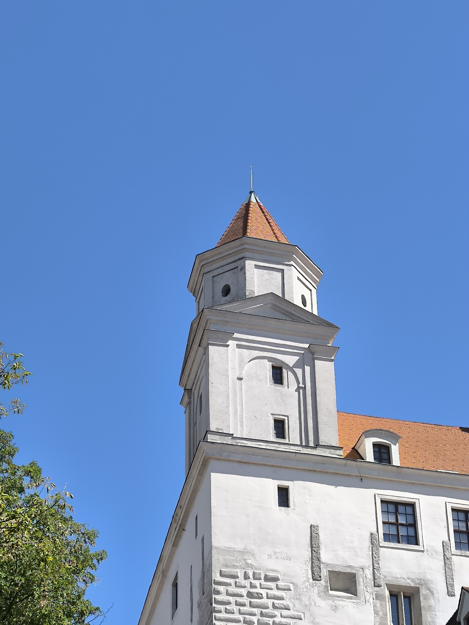 The image shows a close-up of a white tower with a red-tiled roof against a clear blue sky. The tower is part of Bratislava Castle, located in Bratislava, Slovakia. The architecture is historic, with visible stone accents and small windows. A portion of a green tree is visible on the left side of the frame. The scene is static and depicts a bright, sunny day. There are no people or animals visible in the image.