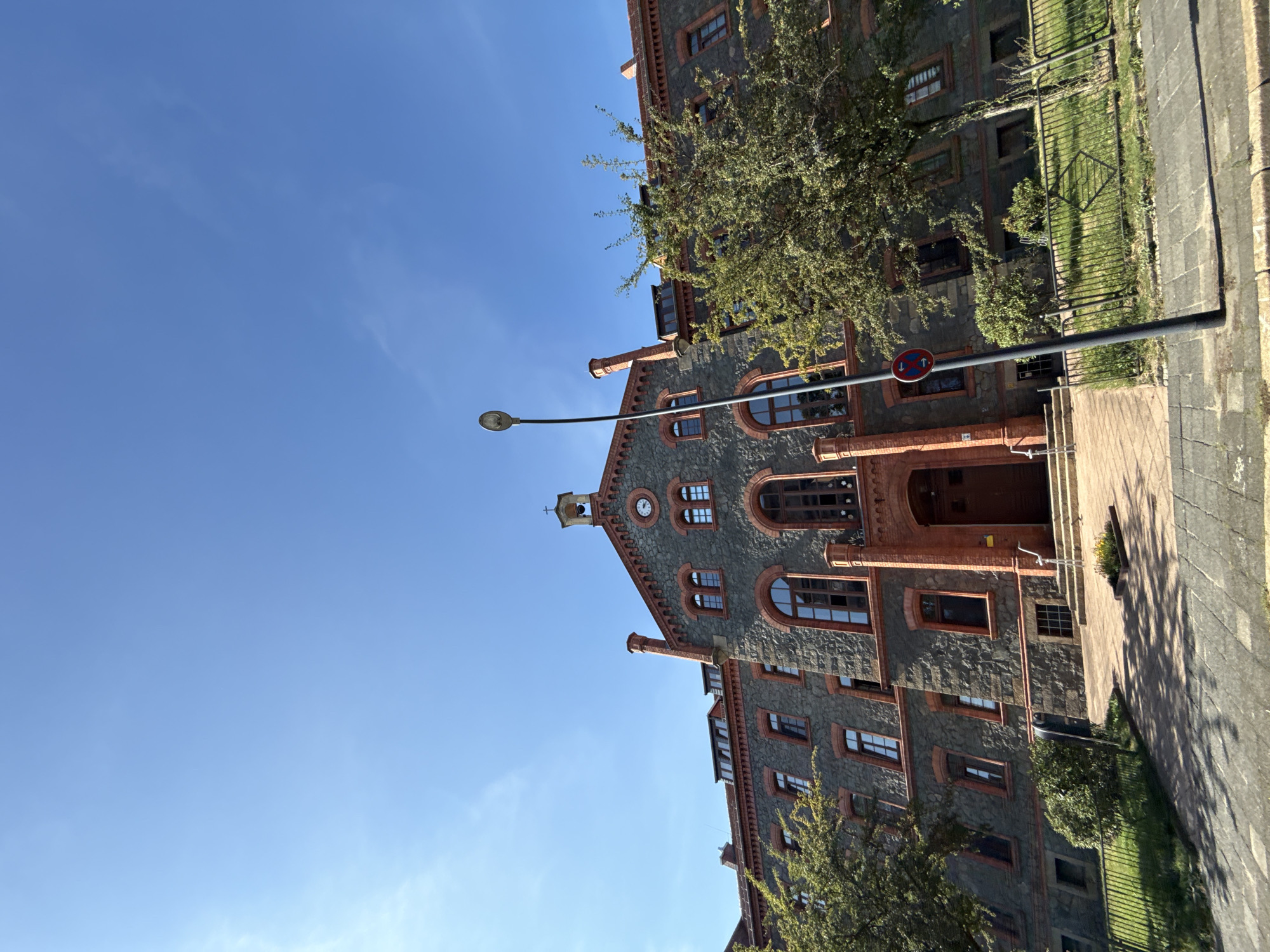 This is a ground-level, wide-angle, vertical photograph taken in the city of Görlitz, Germany, on a clear, sunny day. The main subject is a large, imposing stone building with red brick accents. The building has a dark gray, rough-hewn stone facade, with arched windows framed by red brick. Red brick is also used for the decorative cornices, chimneys, and the entrance portal. The entrance portal is prominent, featuring a wooden door and steps leading up to it. At the apex of the building's roof, a small bell tower with a cross is visible, and below it, a circular clock face is embedded in the facade.
The building is flanked by green trees with dense foliage, some of which are positioned in front of the lower floors. A black lamppost stands to the right of the building's entrance, and a traffic sign showing two red circles with a diagonal line through them is attached to it. In front of the building, there is a paved walkway leading to the entrance steps, and beyond that, a grassy area enclosed by a metal fence. The sky above is a vibrant, clear blue with a few wispy white clouds. The overall impression is that of a historic institutional building, possibly a school or a church, bathed in bright daylight.
