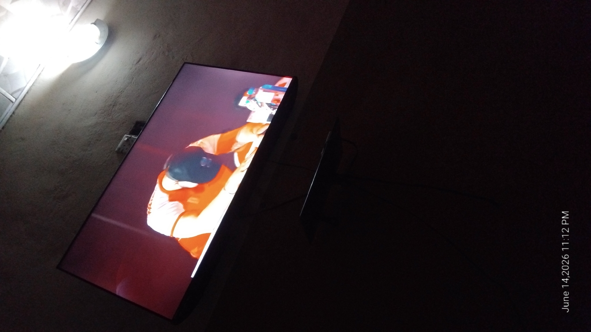 The image depicts a television screen displaying a person wearing an orange and white shirt and a dark baseball cap, with their head down. The television is mounted on a wall in a dimly lit room, with a ceiling light fixture visible in the upper right corner. The wall appears to be textured and painted a neutral color. A dark shelf is mounted below the television. The bottom of the image shows a timestamp: "June 14, 2026 11:12 PM". The scene is consistent with an indoor setting, likely a living room or entertainment area. There is no information in the image to suggest the location within the city of Howayi, Nigeria.