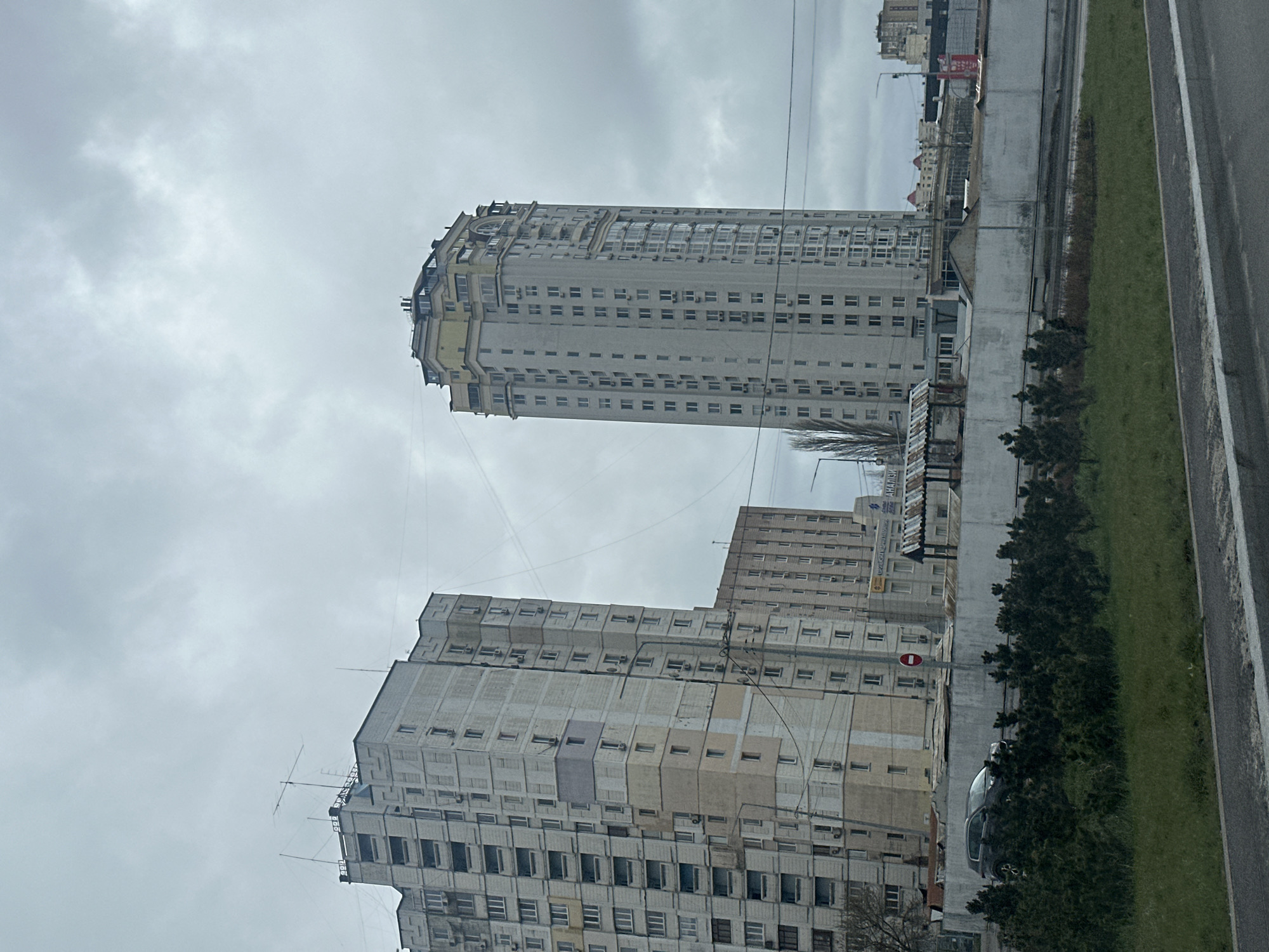 The image shows a street scene in Kyiv, Ukraine, featuring several large, Soviet-era apartment buildings. One tall, slender apartment building dominates the background, while another shorter one sits in front. These are built in a minimalist, utilitarian style and lack decorative elements. The buildings display a grid-like pattern due to the arrangement of windows. In between the shorter apartment block and the road is a car parked on the shoulder, with some small trees and shrubbery in front of the apartment. On the right side of the photo, there is a narrow strip of green grass, a concrete barrier, and part of a street or road. The sky is overcast and grey, suggesting cloudy weather. Overhead wires cross in front of the buildings. The perspective of the photo suggests it was taken from a moving vehicle.