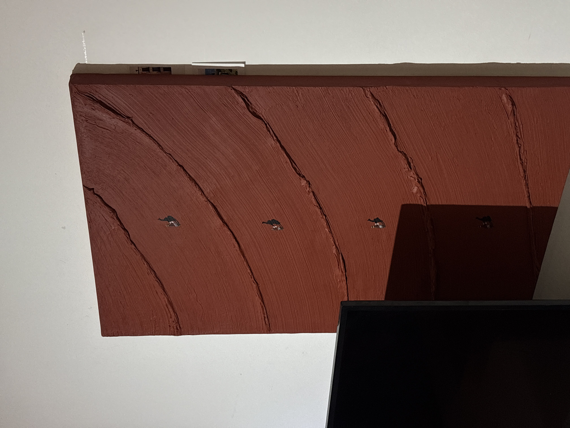 The image shows a portion of a wall in San Bernardino Tlaxcalancingo, Mexico. The wall is painted white, and a reddish-brown, textured art piece is mounted on it. The art piece has a roughly trapezoidal shape with an uneven surface. It appears to be made of plaster or a similar material with vertical striations. Four small, dark figures, possibly representing animals or people, are distributed across the art piece. Above the art piece, a strip of paper or tape with some small images is visible. The lower portion of the image is dominated by a black rectangular shape, likely a television or computer screen. The background is a plain white wall.