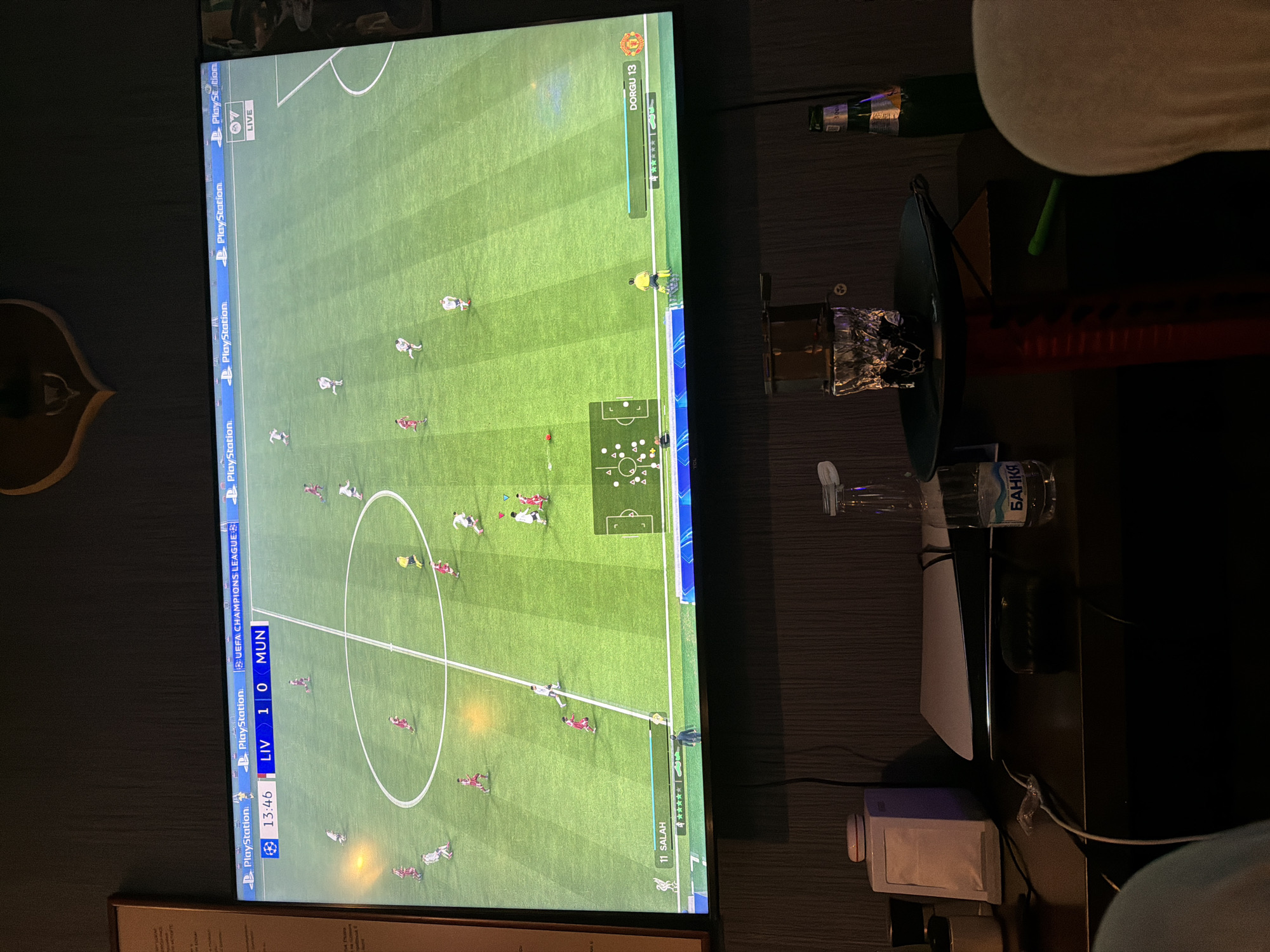 The image captures a dimly lit indoor setting, focusing on a large, widescreen television displaying a football (soccer) video game. The game interface shows an ongoing match between "LIV" (Liverpool) and "MUN" (Manchester United) in the "UEFA CHAMPIONS LEAGUES," with Liverpool leading 1-0 at the in-game time of 13:46. Various "PlayStation" logos are visible along the left side of the screen, indicating the gaming platform. Player names "11 SALAH" and "DORGU 13" are displayed near the bottom and top right of the screen respectively, along with star ratings, suggesting player skill levels. A mini-map of the pitch is also visible in the top right corner of the game screen.
To the right of the television, on a dark surface, a white and black PlayStation 5 console is horizontally positioned. Nearby, a partially visible water bottle with the text "БАНКЯ" (Bankya) is present, along with a small white and blue electronic device, likely a network component or charger, and various cables. A dark, possibly green, bowl or dish containing items wrapped in foil is also on the surface. Another green bottle is barely visible further back on the right. The overall ambiance suggests an evening or nighttime gaming session. Parts of human legs or arms are visible in the top and bottom right corners, indicating the presence of individuals either playing or watching the game.