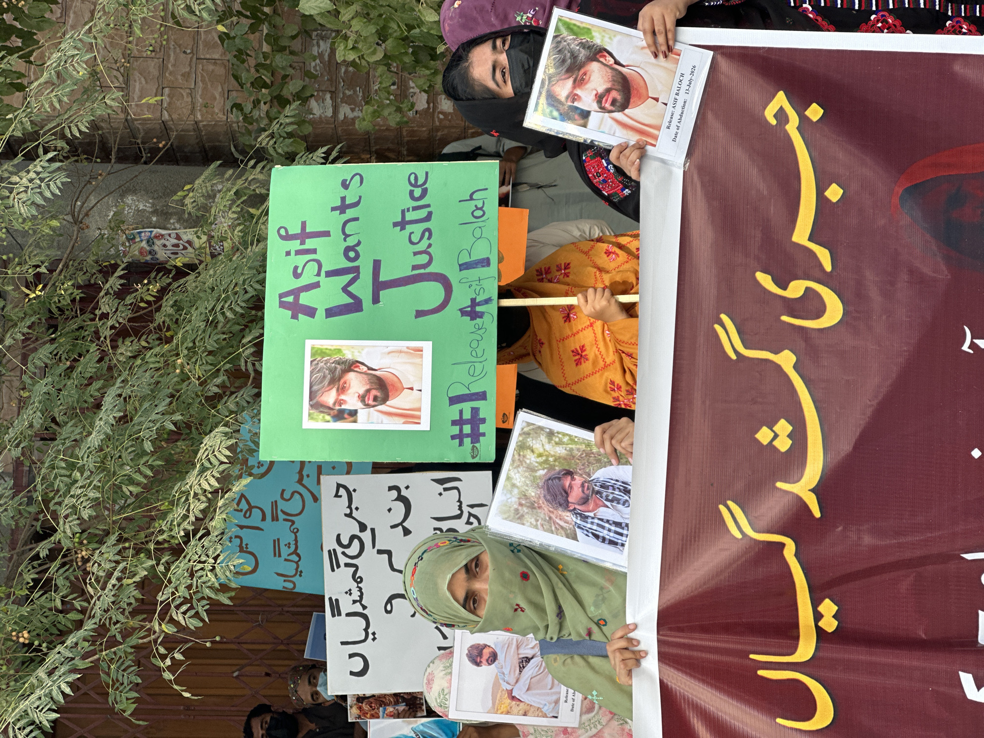 This image depicts a protest taking place outdoors, likely in Quetta City Tehsil, Pakistan, given the script on the signs and banners. In the foreground, three individuals are prominently visible holding signs and photographs. The person on the left, a woman wearing a green hijab that covers her hair and nose, holds a photograph of a man. To her right, another woman, also in traditional attire and wearing a black face mask, holds a photograph of the same man. A third person, partially visible in an orange garment, also holds a sign.
Several signs are displayed. One sign, in English and written in purple marker on a green background, reads "Asif Wants Justice #Release Asif Baloch". Below this, a smaller white label on the photograph held by the woman in black states "Release: ASIF BALOCH Date of Abduction: 13-July-2026". Other signs visible are in Urdu script. One sign, in blue, appears to say "خواتین", meaning "women". Another sign, white with black text, also appears to be in Urdu. A large maroon banner at the bottom of the frame features prominent yellow Urdu text.
The environment suggests an urban setting, with a brick wall and a metal grate visible in the background, along with a tree with green leaves and some small fruit. The overall atmosphere is one of protest and advocacy for the release of someone named Asif Baloch.