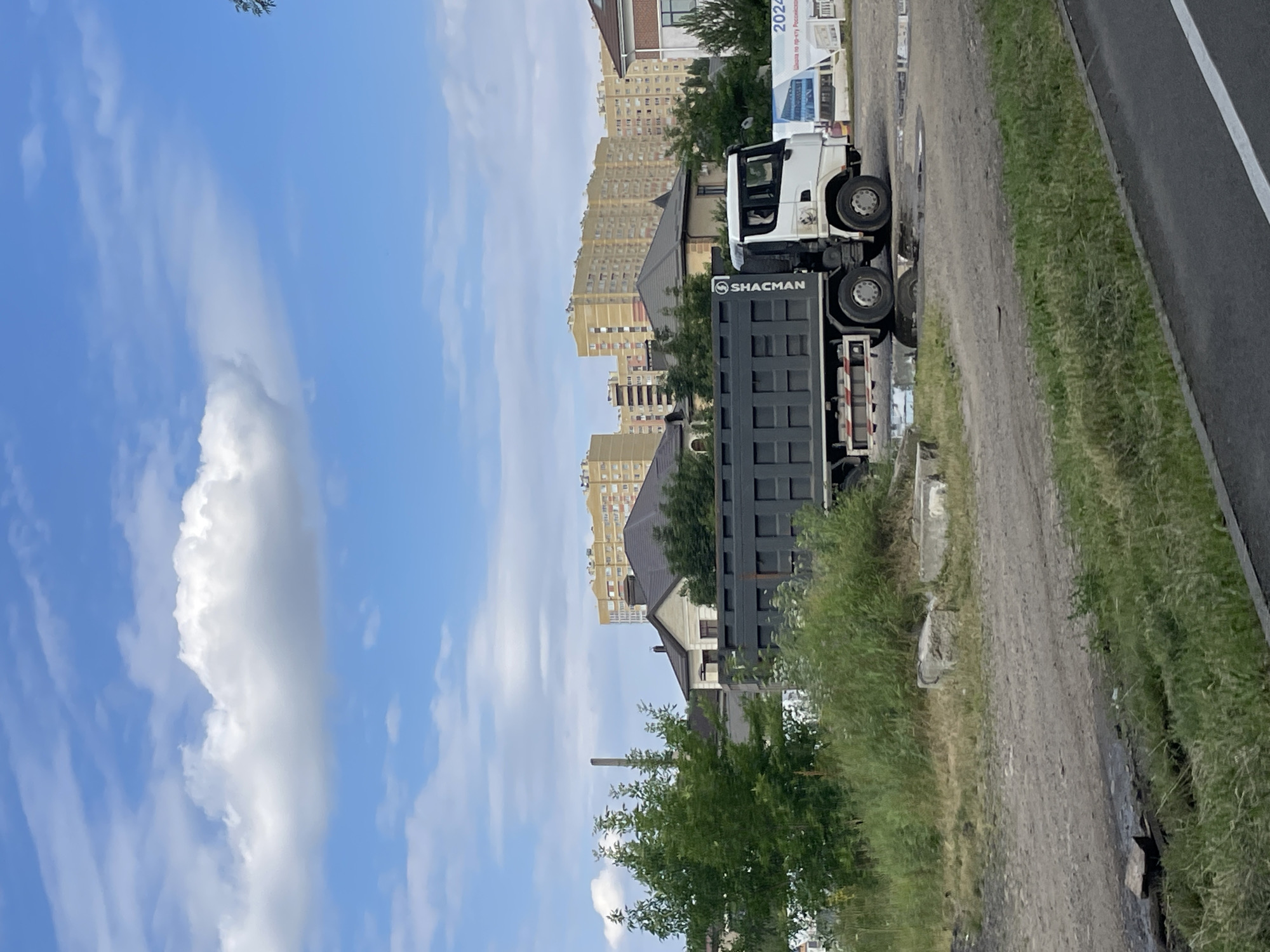 A dump truck with a dark gray bed and a white cab is parked on a dirt and gravel lot. The truck has "SHACMAN" written vertically on its side. Behind the truck, there are several houses with dark roofs and multi-story apartment buildings in yellow and beige. A tall, thin chimney or smokestack is visible between a cluster of trees and a house. A large white banner with blue and red graphics is partially visible to the right of the truck, with the text "2024" and Russian text. The ground is uneven with patches of grass and some puddles of water reflecting the sky. The sky is blue with scattered white clouds.