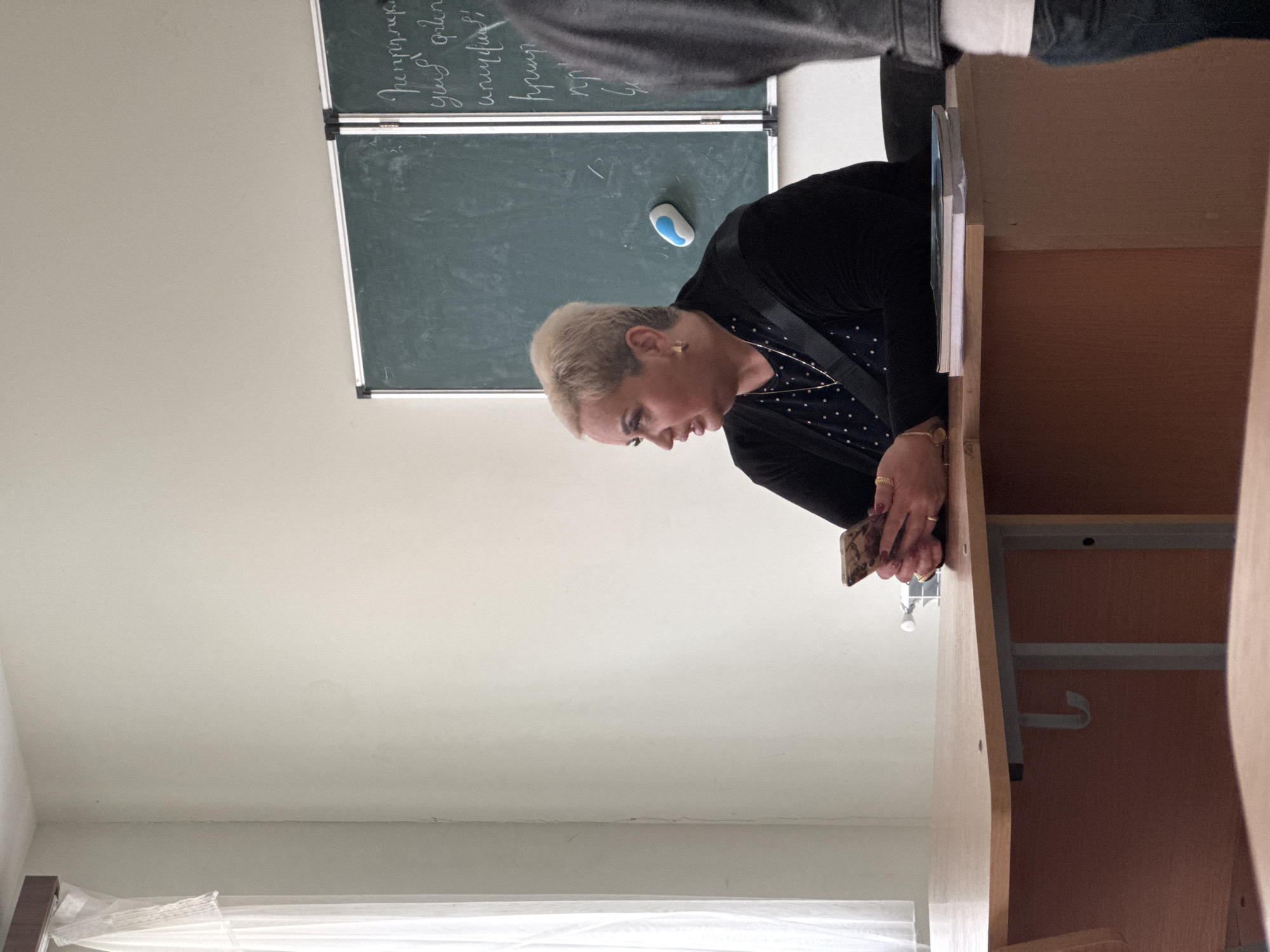 A medium shot, taken in what appears to be a classroom or meeting room in Yerevan, Armenia, captures a person seated at a light wooden desk, engrossed in their smartphone. The person, an adult with short, light blonde hair and fair skin, wears a black long-sleeved top over a black and white polka-dot top, adorned with small gold hoop earrings and a delicate gold necklace. Several rings are visible on their left hand as they hold the smartphone, which has a patterned case.
Behind the person, on the left side of the frame, are two dark green chalkboards, stacked vertically. The upper board features writing in Armenian script, including words such as "Պետերբուրգ" (Petersburg), "գործուն" (active/effective), "գյուղ" (village), "քաղաք" (city), "որպես" (as), and "հրապարակ" (square/public place). The lower board has less distinct writing, with what appears to be a letter and perhaps a number "2". A blue and white chalk holder or eraser rests on the bottom blackboard.
On the desk to the right of the seated person, a stack of notebooks or a book is visible. A window with white sheer curtains or blinds is partially seen in the lower left corner, indicating daytime. Another person, seen from the back, wearing a dark top, is partially visible in the upper right corner of the frame. The overall atmosphere suggests a casual moment during or between formal activities.