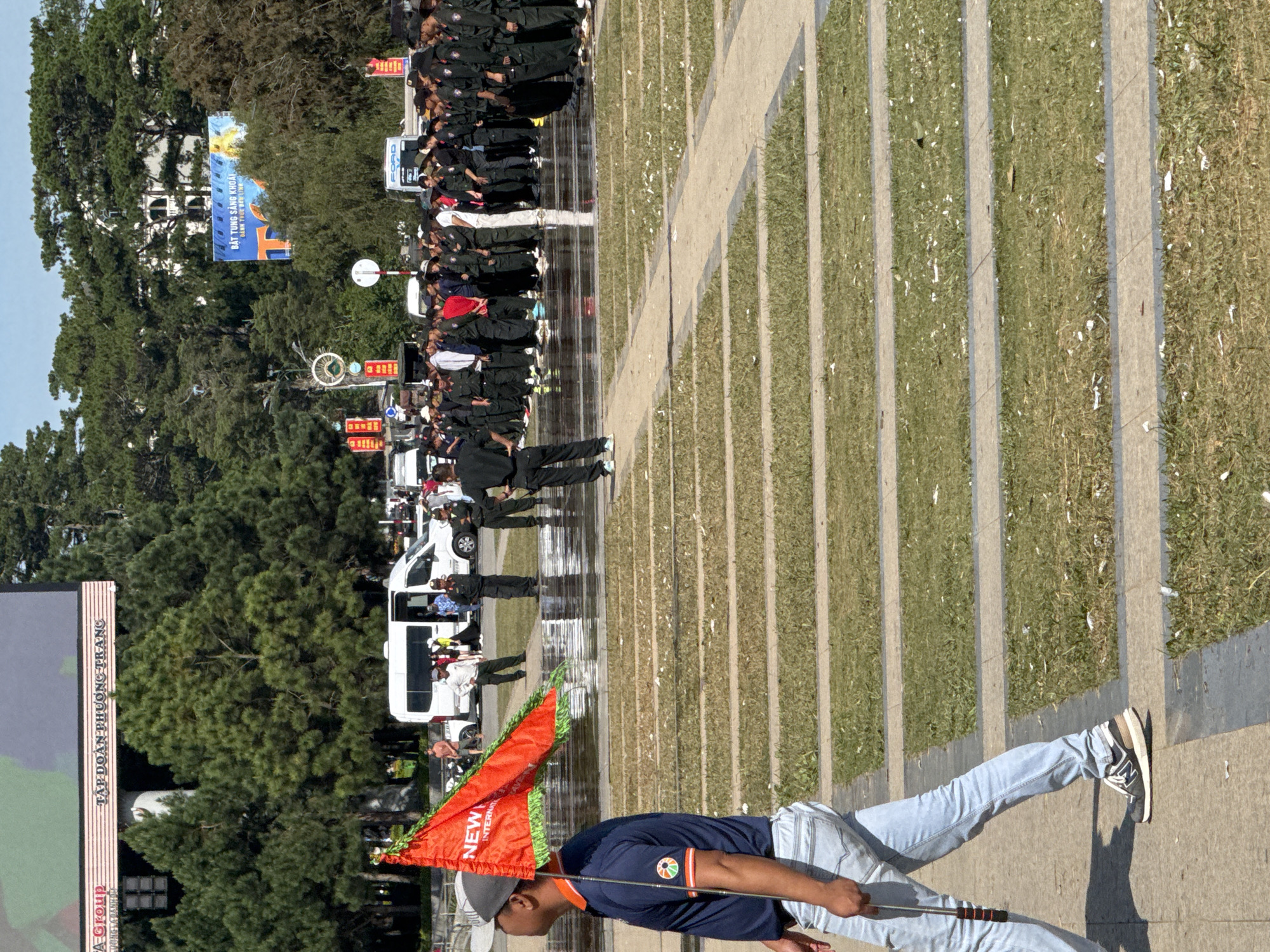 In Da Lat, Vietnam, a man in a blue polo shirt and light-wash jeans walks past a reflective wet ground, holding a red flag with green trim. The flag displays the word "NEW" in white letters. Behind him, a line of people in dark uniforms stand at attention, some holding what appear to be flags or poles. In the background, several white vans are parked, and lush green pine trees frame the scene. Large billboards are visible above the trees. The overall environment suggests an outdoor public space or event.