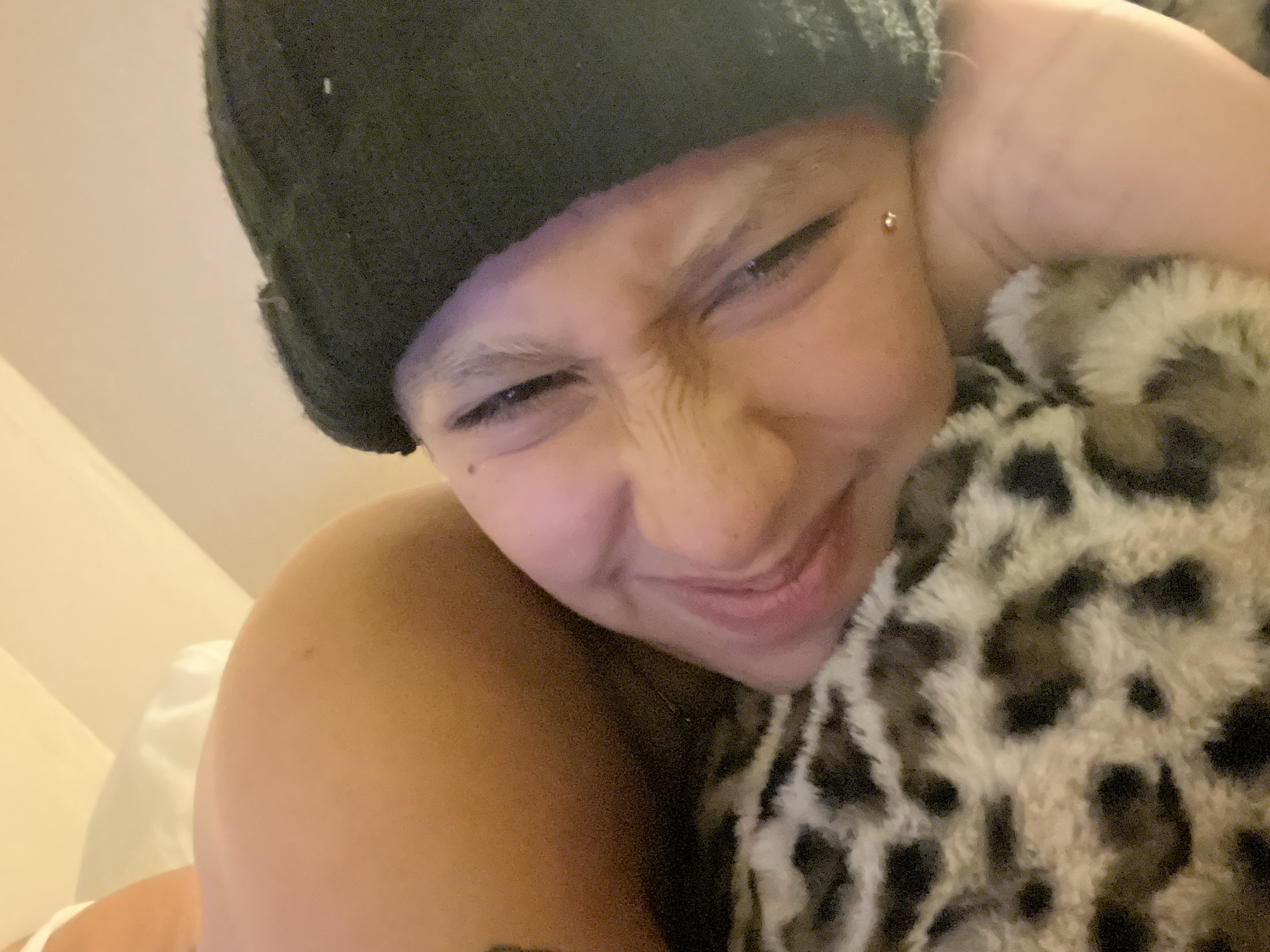 A close-up of a person wearing a black beanie with short hair, a helix piercing above the left eye, embracing a stuffed animal with a leopard print. The background appears to be a bedroom with a white pillowcase visible. The person has their eyes squinted and appears to be smiling. The location is not discernible from the image.
