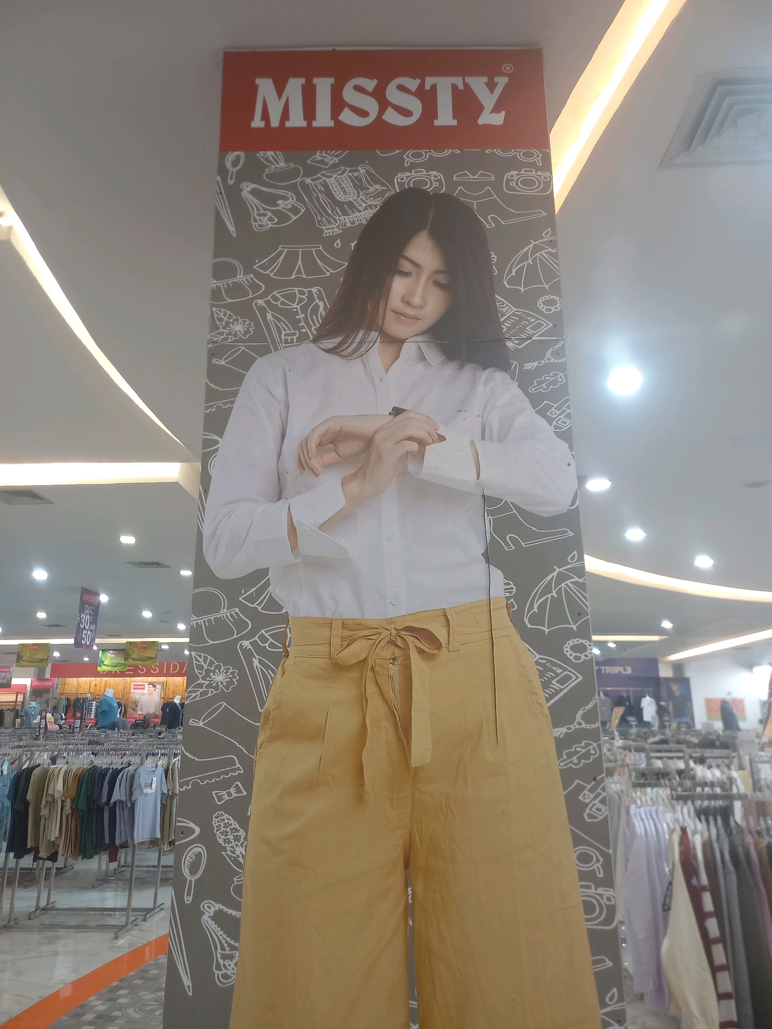 A young woman is standing in front of a large poster with "MISSTY" written in large red letters at the top. She is wearing a white button-down shirt with rolled-up sleeves and yellow wide-leg pants. Her hands are clasped in front of her, and she is looking down at her left wrist. The poster behind her has a pattern of line drawings of clothing and accessories. The background appears to be a retail environment with clothing racks and displays visible. The lighting suggests an indoor shopping mall or store. There are no other people clearly visible in the immediate vicinity. The scene is likely in Tunggorono, Indonesia, given the information provided.