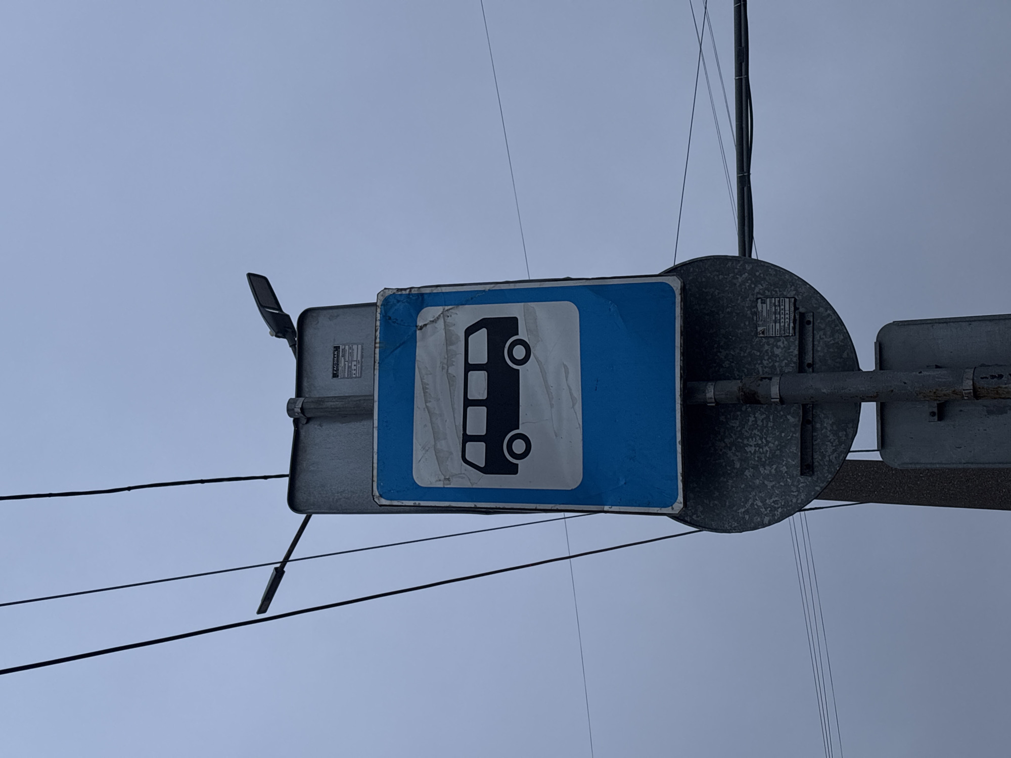 The image shows a weathered blue and white bus stop sign in Perm, Russia. The sign depicts a stylized bus and is mounted on a metal pole, with various electrical wires and components visible around it. The sky is overcast and gray.