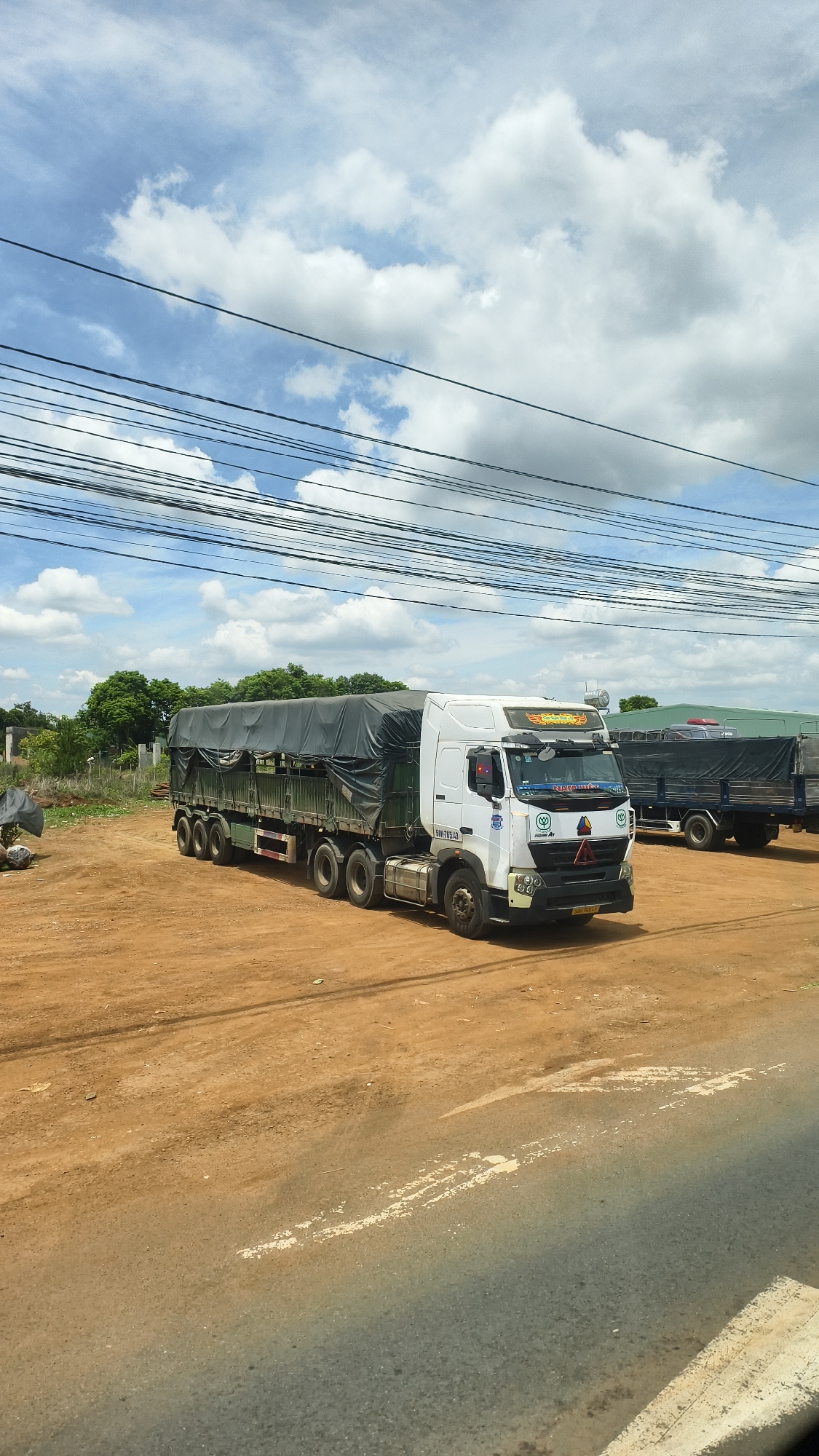 A white semi-trailer truck with a dark tarp-covered cargo bed is parked on a dirt lot under a cloudy blue sky in Chư Sê, Vietnam. The truck has a white cab with a logo on the windshield and a license plate that reads "59H-705.63". Another semi-trailer truck, mostly obscured, is parked behind it. The lot is covered in reddish-brown dirt and gravel, with some sparse vegetation and trees in the background. Overhead, a network of black power lines crisscrosses the frame. The scene appears to be a rest stop or a staging area for trucks.