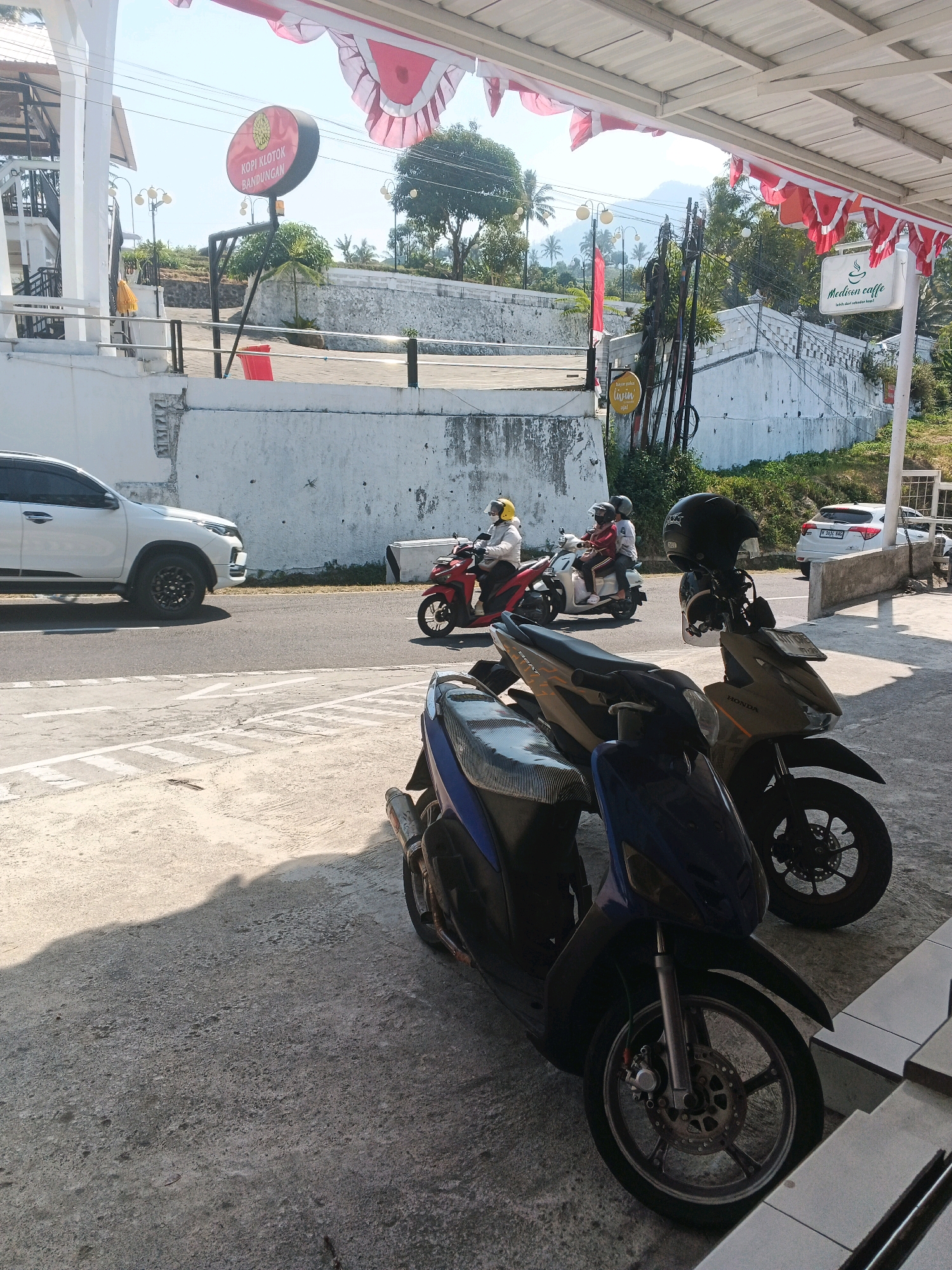 The image captures a street scene in Pakopen, Indonesia. In the foreground, two scooters are parked on a concrete surface. One is blue and black, and the other is a sandy gold color with a black seat. A black helmet rests on the handlebars of the gold scooter. In the middle ground, a white SUV is parked on the side of a paved road. Several people are riding motorcycles on the road. Two individuals on a red scooter are visible, followed by two people on a cream-colored scooter. A white sign with red lettering that says "Melliven caffe" is attached to a white pole. A circular sign with red and yellow colors is also visible. Red and white flags are strung along the edge of an overhang. In the background, there are white walls, trees, and hills under a clear blue sky.