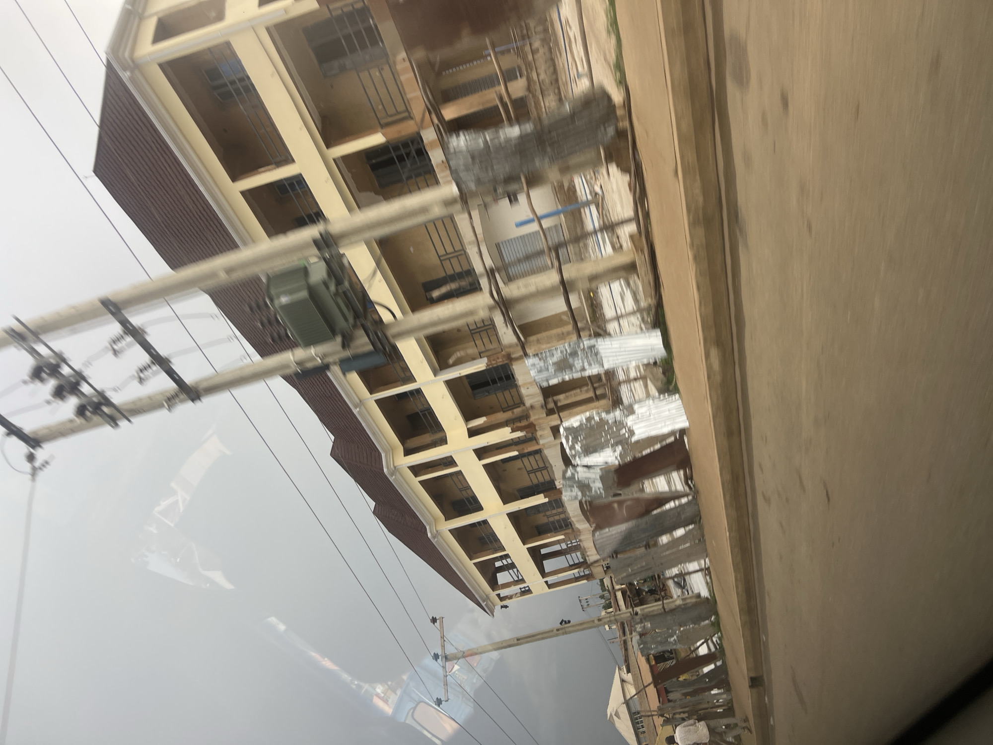 The image shows a multi-story building under construction in Bukuru, Nigeria. The building is painted yellow with white trim and appears to have multiple balconies or open window frames. Some sections of the building are covered with corrugated metal sheeting, suggesting ongoing construction work.
In the foreground, there are power lines and a utility pole with some electrical equipment mounted on it. The power lines extend across the scene and are a prominent feature. To the right of the building, a concrete road or sidewalk is visible, with a curb separating it from the construction area.
The overall impression is that of a developing urban or suburban environment. The sky is overcast, indicating a cloudy or possibly rainy day. There are no visible people in the immediate foreground, but some figures might be present in the distance near the construction site, though they are not clearly discernible.
The image is taken from a slightly elevated perspective, possibly from a moving vehicle, as indicated by the angle and slight blur in some areas. The scene appears to be captured during daylight hours, but the cloudy weather might make it difficult to determine the exact time of day. There is no visible text in the image.