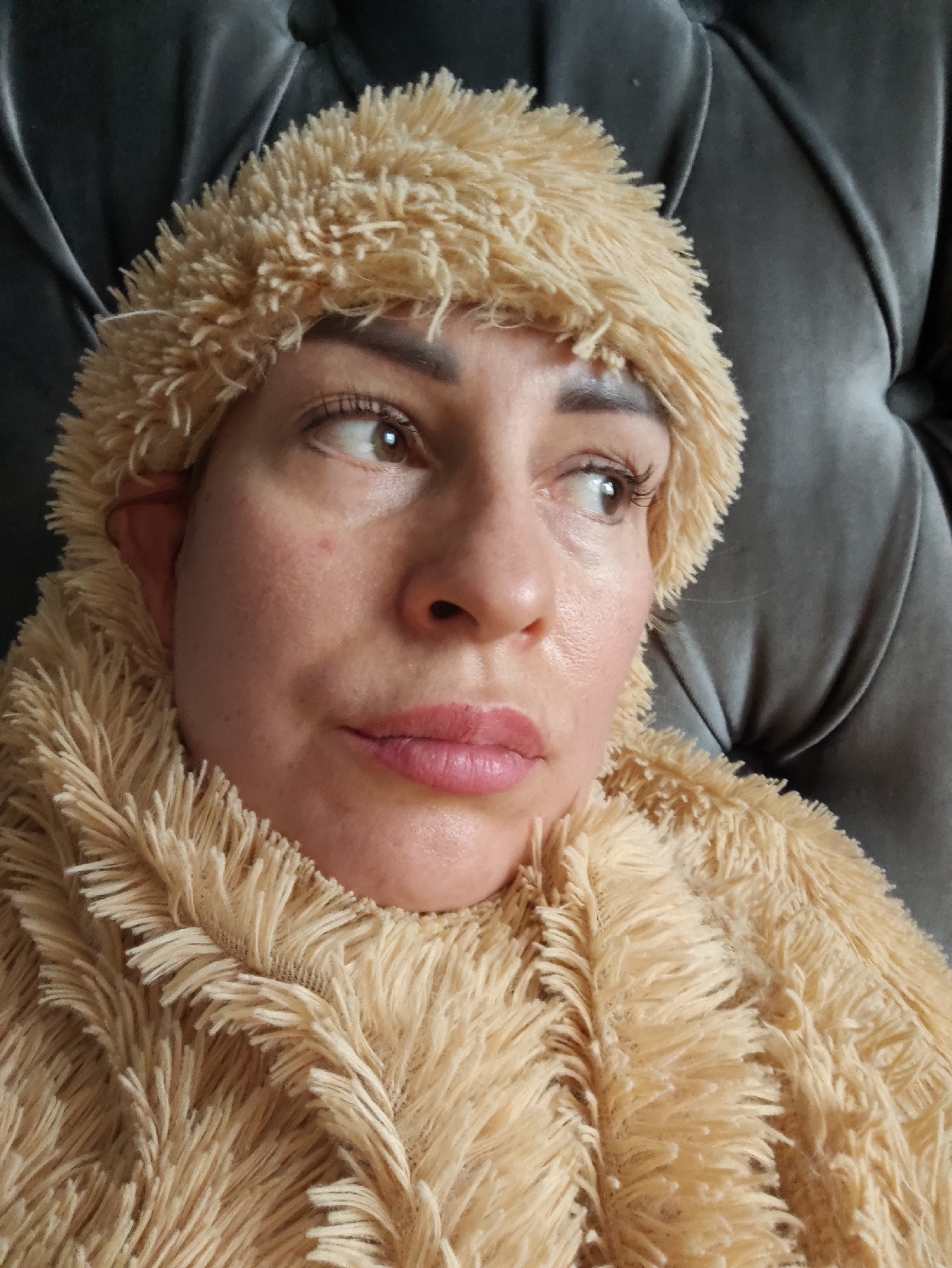 This is a close-up, eye-level shot of a woman wearing a tan, fuzzy, hooded garment that covers her head and neck. Her eyes are looking off to the right, and her lips are slightly parted. Her skin appears somewhat oily, and there are a few blemishes visible on her cheek. The background is a dark, textured surface, possibly a sofa. The lighting is soft and appears to be coming from the front. The overall mood conveyed is one of introspection or perhaps weariness. There is no visible text in the image.