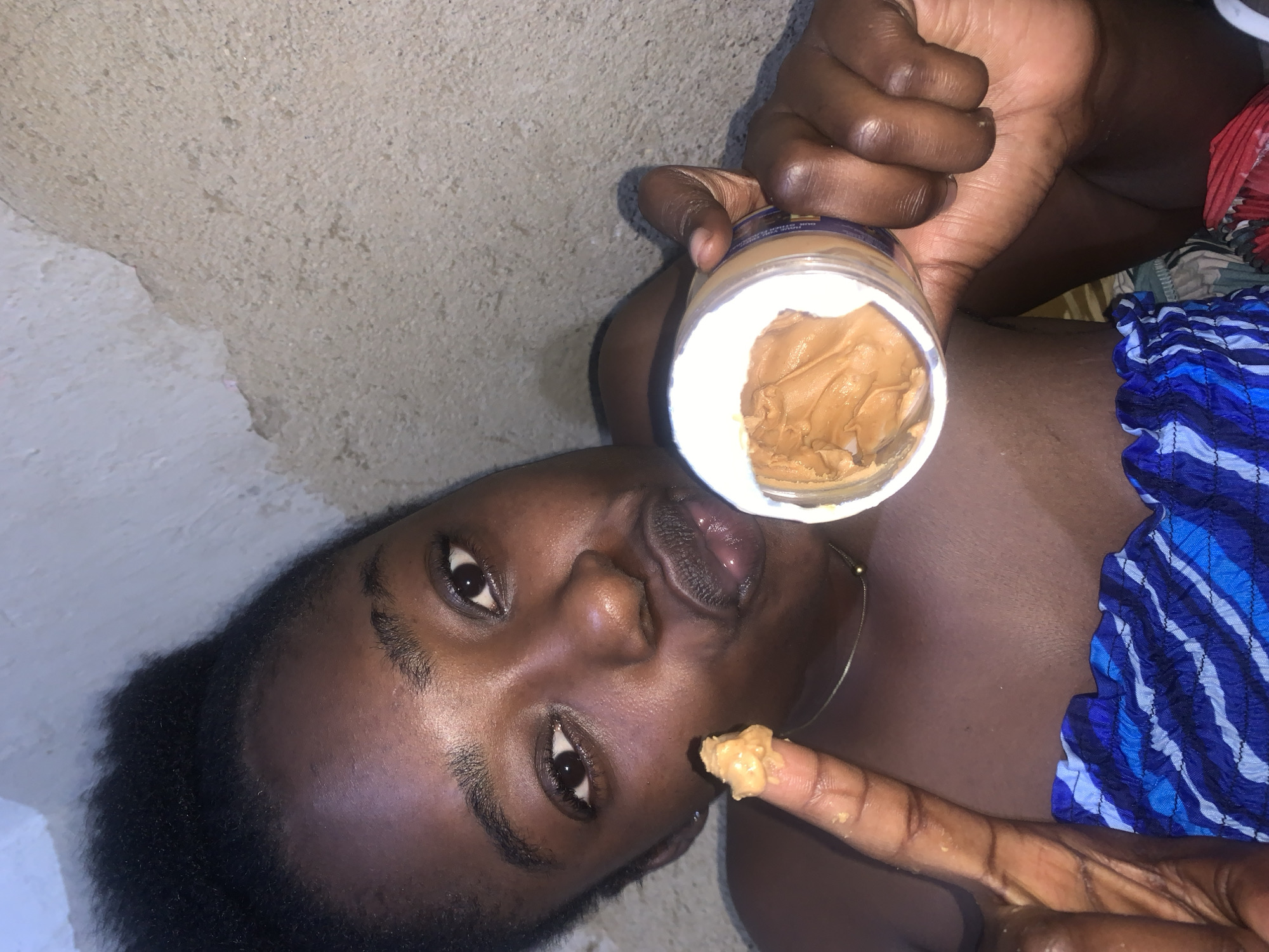 The image shows a young Black woman in what appears to be an indoor setting with rough, textured walls in the background. She is holding a jar of peanut butter and has some on her index finger, which she is bringing towards her mouth. Her mouth is slightly open in a puckered expression. She is wearing a blue and white striped top. The scene does not contain any information that would allow for a specific location in Rafin Par, Nigeria to be identified.