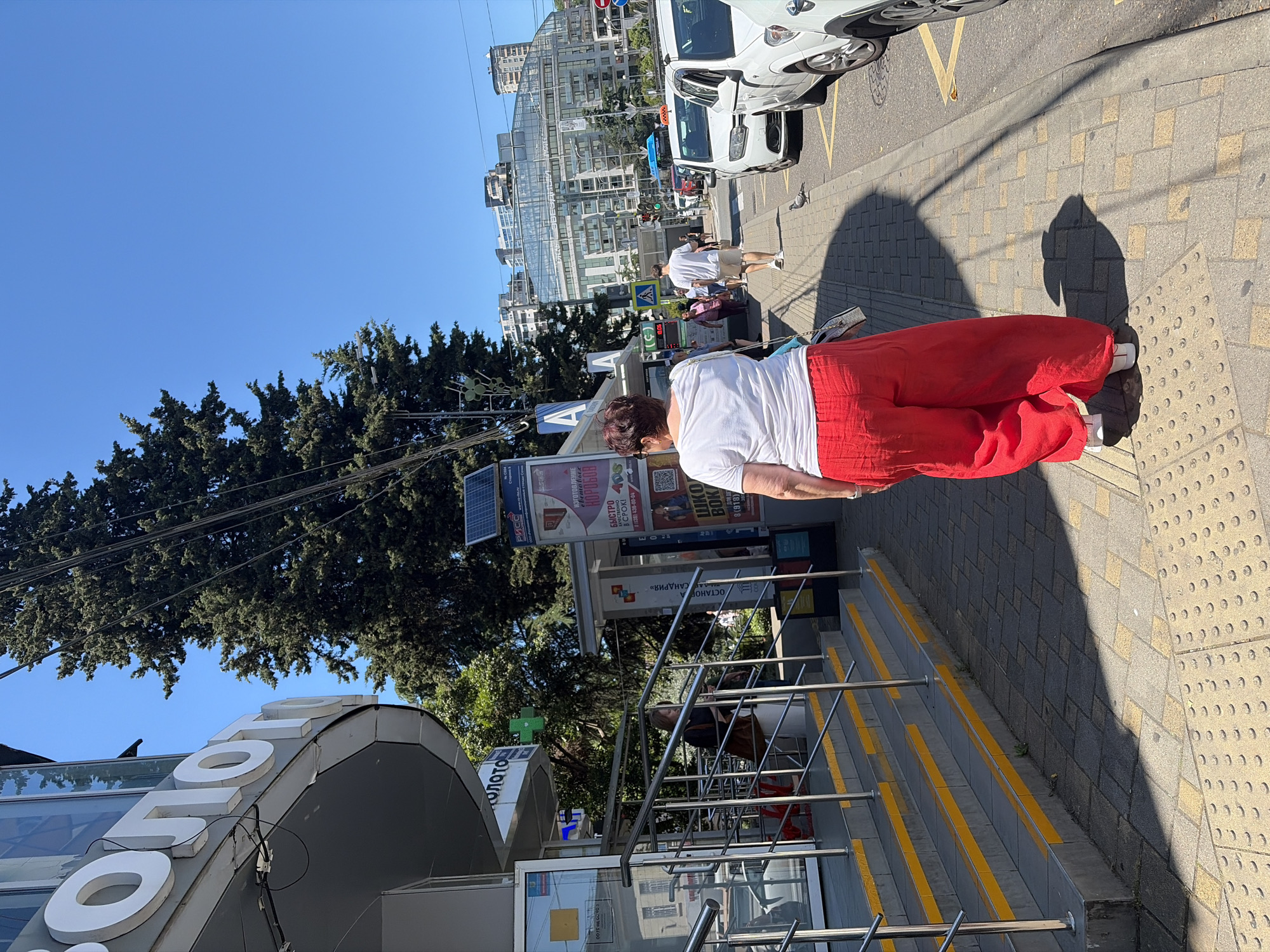 An outdoor, medium-shot, eye-level, full-body photograph captures a woman walking on a sidewalk in Sochi, Russia. The woman, with her back to the camera, is wearing a white t-shirt and flowing red pants. She carries a small silver purse on her left shoulder. The sidewalk is paved with interlocking square and rectangular tiles, with a row of tactile paving circles running along its edge. Sunlight casts a long shadow of the woman on the ground.
To the left of the sidewalk, a staircase with yellow-lined steps ascends to an entrance of a building. The building has a curved facade with large white letters spelling "ЗОЛОТО" (ZOLOTO, meaning "gold"). Above the entrance, a green cross, a common symbol for a pharmacy, is visible. Further down the street, other buildings, including a modern glass-fronted structure, line the horizon. Cars, including white taxis, are parked along the street, which has yellow lane markings. A pigeon is visible on the sidewalk in the middle ground. The sky is clear and blue. The overall scene depicts a sunny day in an urban environment.