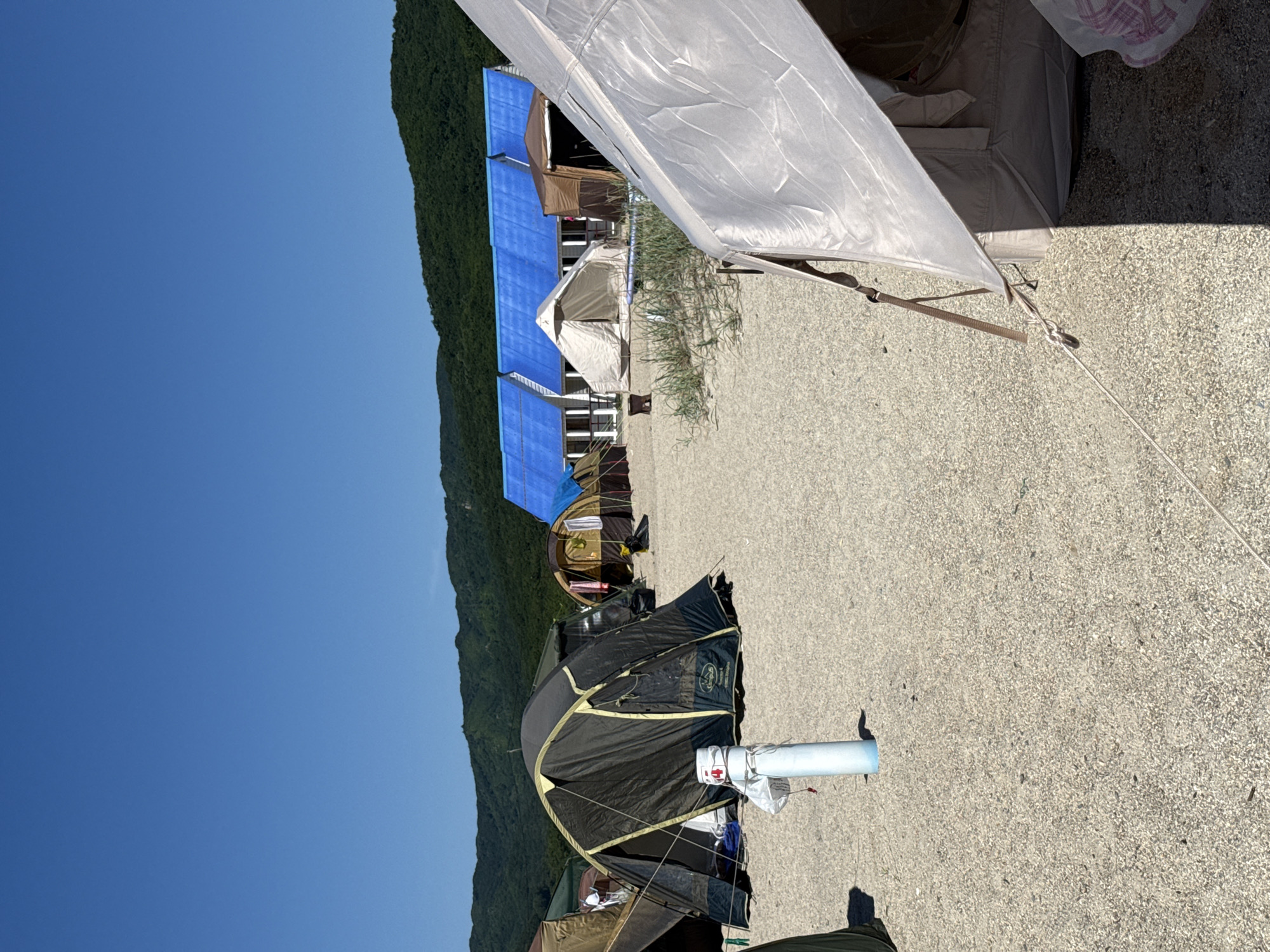 The image depicts a campground scene under a clear blue sky. In the foreground, a light-colored tent is partially visible, its fabric casting a shadow on the sandy ground. A white cylindrical object with blue at its base stands near the center of the frame, with a white bag and some rope attached to it. Behind this, a large, dark-colored tent with a yellow trim is pitched. Further in the background, several other tents of various colors and sizes are set up, including a group of tents with blue tarps covering their roofs. Beyond the tents, a row of buildings with white walls and blue roofs is visible, and behind them, a densely forested hill rises under the sky. The ground is covered in small pebbles or gravel. There are no people visible in the image. The location is identified as Nakhodka, Russia.