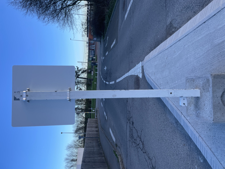 The image displays a street scene in Plaisir, France, viewed from directly behind a plain white, rectangular road sign. This sign is affixed to a white metal pole via two silver-colored clamps. The pole is anchored within a triangular, light grey concrete traffic island that protrudes into the road in the foreground.
The road surface is dark asphalt, exhibiting visible cracking, particularly on the left side of the frame. White painted markings delineate the traffic flow: a solid line bordering the concrete island, dashed lines indicating lanes, and two distinct arrows on the pavement suggesting a left turn and a straight-ahead direction.
To the left, a tall, weathered wooden fence follows the curvature of the road. Beyond the fence, a light-colored building and numerous deciduous trees with bare branches are visible. A streetlight pole stands further down the road on the left. On the right side of the road, a patch of natural ground with similar leafless trees and several white bollards are present.
In the background, a multi-story building with reddish and white sections, potentially a station, is visible. Overhead wires and a bridge-like structure, characteristic of a railway line, are discernible above and behind this building. A solitary, indistinct individual is present in the distant background near the building on the right. The sky above is clear and blue, indicating natural daylight conditions.