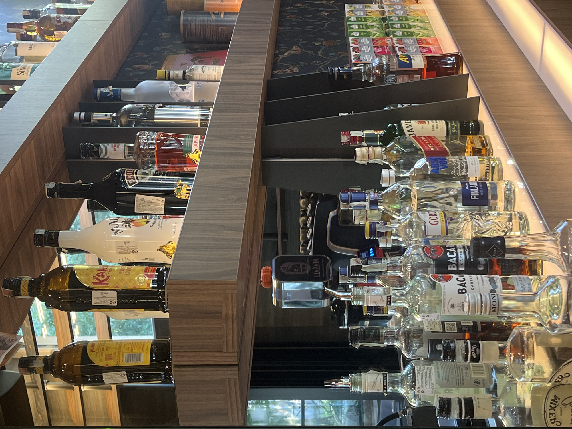 This image depicts a well-stocked bar or liquor store shelf filled with numerous bottles of alcohol. The bottles are arranged in rows on dark shelves, with a reflective surface below them that mirrors the products. The scene is brightly lit, suggesting an indoor environment, possibly in the evening or with artificial lighting.
The bottles include a variety of spirits, such as vodka, whiskey, and liqueurs, with labels from recognizable brands like Absolut, Finland, Bacardi, and Johnnie Walker (Red Label). There are also some less common or specialty bottles, indicated by their unique shapes and labels. Some bottles are clear, showcasing the liquid inside, while others are colored glass.
The shelf unit is angled, creating a dynamic perspective. A patterned wallpaper or fabric is visible behind some of the bottles on the upper shelves, adding a decorative element to the setting. The overall impression is one of abundance and variety, typical of a place dedicated to the sale or serving of alcoholic beverages. No people are visible in the image.