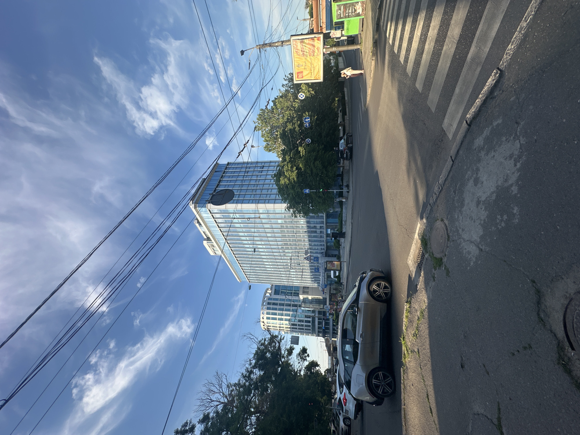This image captures a street scene in Odesa, Ukraine, on a clear, sunny day. The perspective is low and tilted, giving a dynamic feel to the urban landscape. Dominating the background are two modern glass-fronted buildings, reflecting the bright blue sky with scattered wispy clouds. In the foreground, a pedestrian crosswalk is clearly visible on the asphalt road.
A silver electric car, possibly a BMW i3, is parked on the right side of the street, its futuristic design contrasting with the more traditional elements of the scene. A person is walking across the crosswalk in the distance, and another individual appears to be standing on the sidewalk near a bus stop. Overhead, a complex network of power lines crisscrosses the sky, a common sight in many urban environments.
A large billboard with text in a foreign language is prominently displayed on a pole, suggesting commercial activity. Various street signs and traffic lights are also visible, indicating a functioning urban thoroughfare. The overall impression is one of a contemporary city, bustling with everyday life under a pleasant sky. The sunlight casts long shadows, suggesting it is either morning or late afternoon.