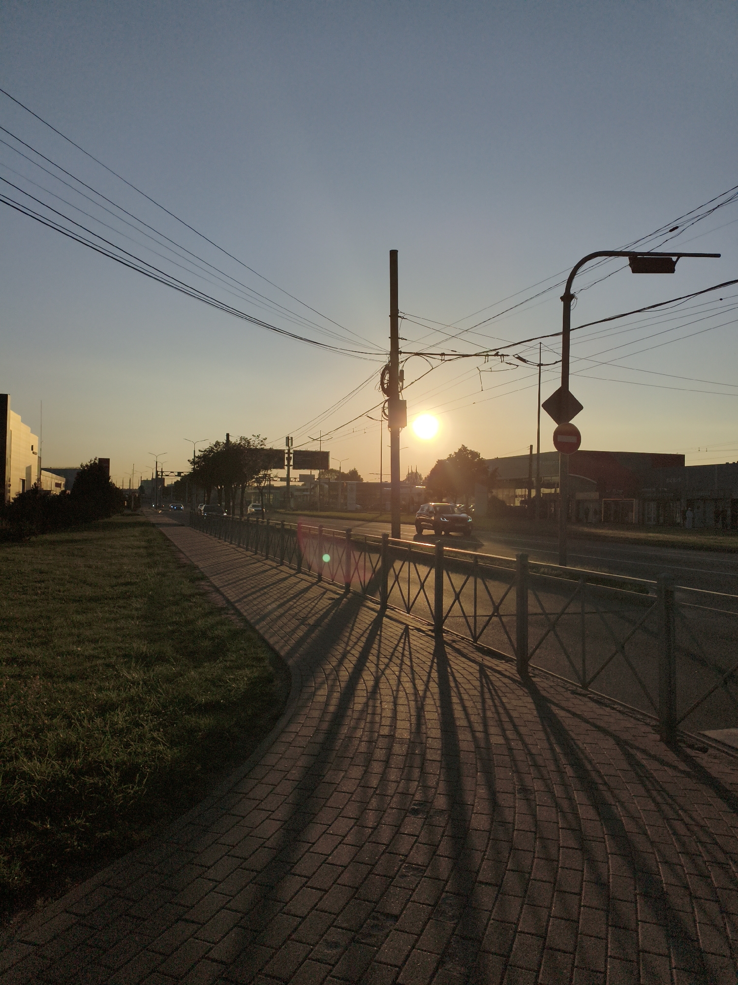 An eye-level, medium shot captures a street scene at sunset. The sun, a bright orb, is positioned in the upper center-right of the frame, casting long shadows from a metal fence onto a brick sidewalk. The sidewalk curves away from the viewer, bordering a strip of green grass on the left. In the background, a two-lane road with several cars is visible, with buildings and streetlights lining the opposite side. Overhead, a network of power lines crisscrosses the sky, silhouetted against the pale orange and blue gradient of the twilight sky. A traffic light and two traffic signs are mounted on a pole near the road. The overall atmosphere is serene, with the setting sun illuminating the urban landscape.