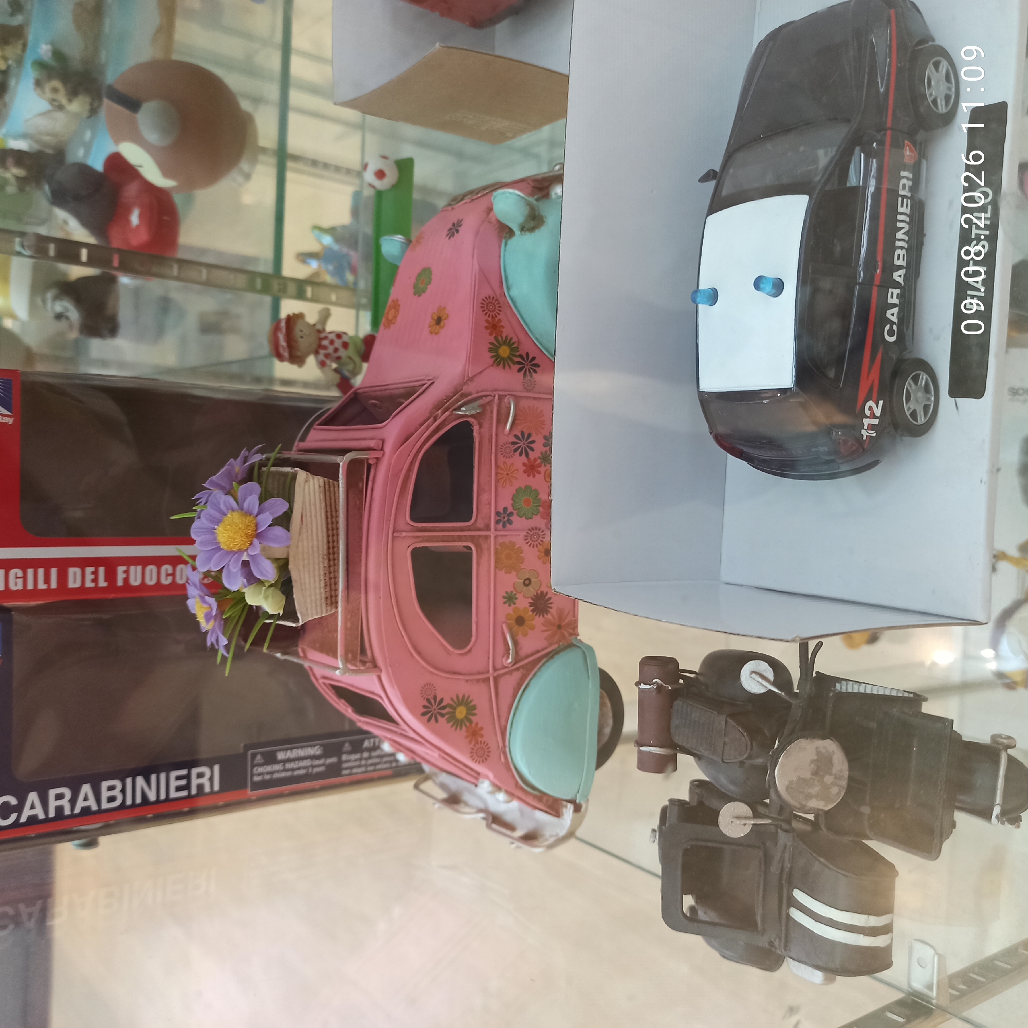 A glass display case contains several collectible toys. In the foreground, on the left, is a dark-colored toy motorcycle with a sidecar, decorated with two white stripes. To the right of the motorcycle is a black toy car with a white roof, blue lights, and "CARABINIERI" written on its side. The car has the number "112" on its rear. Behind these toys, a pink vintage-style toy car with a floral design is displayed. On the roof of the pink car is a small basket with artificial purple and yellow flowers. Above and behind the pink car, a red and white box is visible with "CARABINIERI" and "VIGILI DEL FUOCO" printed on it. The display case appears to be located in a shop or museum in Molfetta, Italy, as indicated by the timestamp "09.08.2026 11:09" on the black toy car, which likely includes a date and time stamp.
