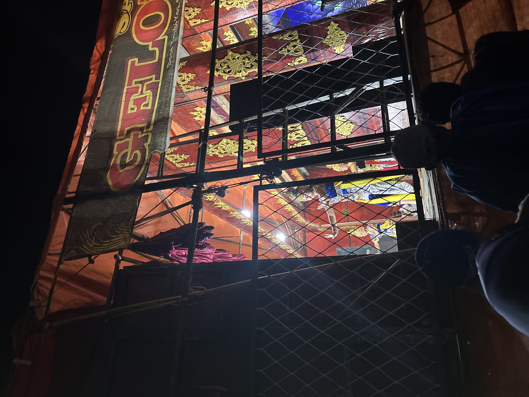 The image depicts an outdoor cultural performance, likely a traditional dance or play, taking place at night in Panongan, Indonesia. The stage is set up under a large orange tarp, with a dark banner featuring red and yellow lettering that appears to read "GET LOH" prominently displayed above the performance area. The stage backdrop is decorated with ornate red and gold patterns.
Visible on the stage are at least two performers dressed in elaborate, colorful costumes and traditional headwear. One performer, in the center, is captured mid-pose with their hands outstretched. Another performer is visible to their left. The stage is lit by bright lights, illuminating the performers and the decorative backdrop.
In the foreground, a wire mesh fence separates the audience from the performance area. Several audience members, mostly seen from behind, are gathered to watch. Their silhouettes are dark against the illuminated stage, with some wearing hats and casual clothing. The overall atmosphere suggests a community event or local festival. The setting appears to be a temporary stage erected for the occasion, likely in an open area within the city.