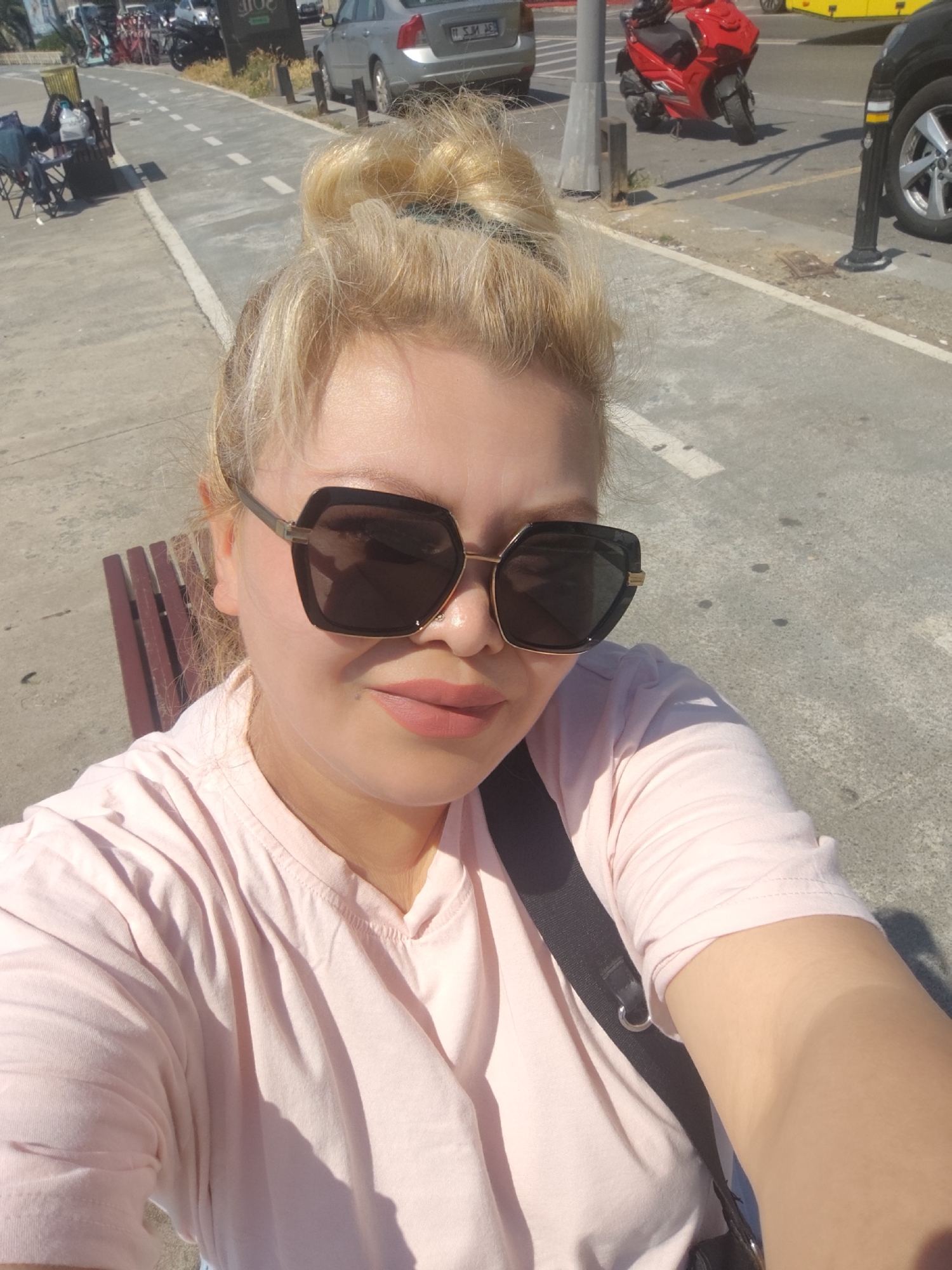A woman is taking a selfie outdoors in Sarıyer, Turkey. She has blonde hair styled in a bun and is wearing large sunglasses and a light pink T-shirt. A black strap crosses her shoulder. She is sitting on a bench.
In the background, there's a street with parked cars, scooters, and bicycles. A designated bike lane is visible with white dashed lines. Other urban elements include lampposts and street signs. The photo appears to have been taken during the daytime, as the lighting is bright.