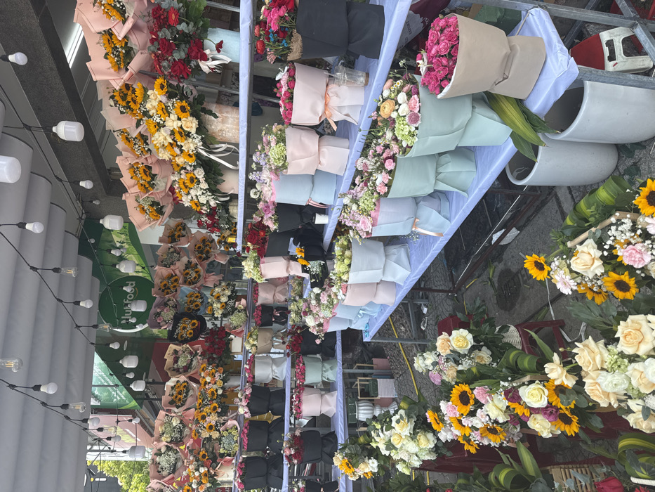 This image captures an outdoor flower stall in Ngũ Hành Sơn Ward, Vietnam, displaying a vast array of pre-arranged bouquets on multi-tiered metal shelving units covered with light-colored fabric. Overhead, a string of bare Edison-style and white spherical light bulbs provides illumination, with a green "clubfood" sign visible in the background. The stall features numerous bouquets, prominently featuring sunflowers mixed with white or yellow roses and filler flowers, often wrapped in peach, pink, light blue, or black paper. Other arrangements include various roses (red, pink, white, yellow), carnations, hydrangeas, and gerbera daisies, wrapped in pastel shades like light pink, light blue, mint green, beige, and black. Some flowers are displayed in white ceramic-like vases or woven baskets on lower levels. The ground is paved, and small red stools along with white cylindrical planters are visible near the base of the display. No people are present in the scene.