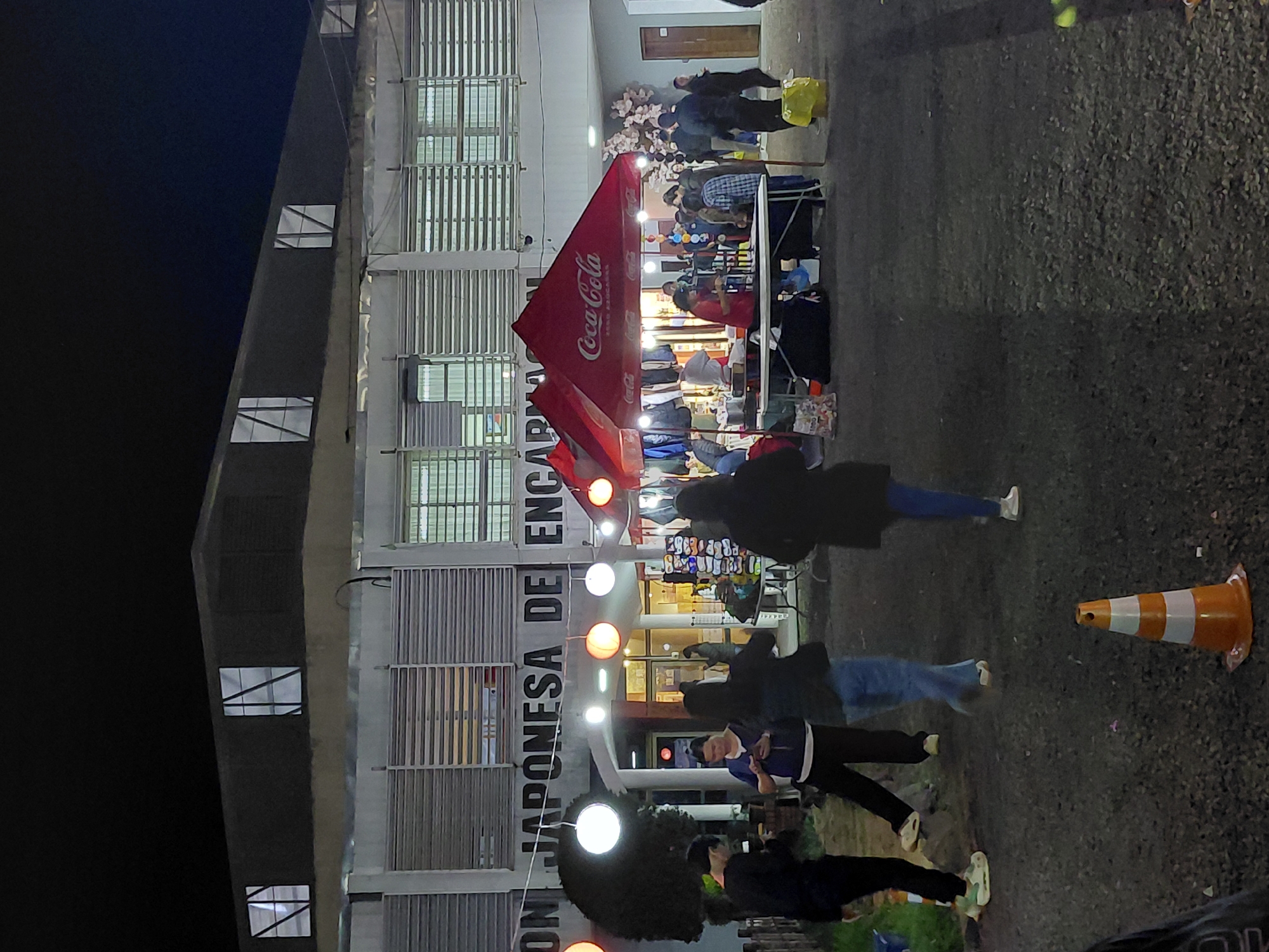 The image depicts an outdoor night market in Encarnación, Paraguay, likely held in front of a community center or municipal building. The architecture suggests a functional, possibly public, structure with horizontal louvers on the upper facade and large windows. In the foreground, several individuals are walking or standing, engaged in various activities typical of a market setting. A red Coca-Cola branded tent is prominently displayed, suggesting a vendor area. Various stalls and displays are visible behind the tent, with items for sale, including clothing. Decorative lanterns, some spherical and lit, are hung, indicating a festive atmosphere. A traffic cone is present in the lower right quadrant, and a yellow trash bag is visible near the Coca-Cola tent. The ground appears to be paved with gravel. The overall scene captures a lively, public event in a South American urban environment at night.