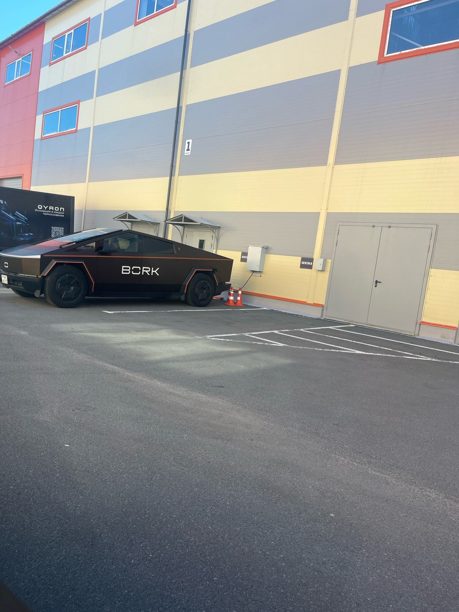 The image captures a side view of a dark-colored electric vehicle, resembling a Tesla Cybertruck, parked in an asphalt lot adjacent to a multi-story industrial building. The building features horizontal bands of cream, light gray, and darker gray on its upper sections, with red-framed windows. On the left side of the building, a section is painted pink. The vehicle has the word "BORK" written in white on its side. Orange trim accents the vehicle's design. Two orange traffic cones are visible in front of the vehicle. The lot has white painted lines, possibly indicating parking spaces. In the background, a large gray double door is set into the building. The overall scene suggests an outdoor exhibition or display area, possibly at a commercial or industrial facility in Saint Petersburg, Russia.
