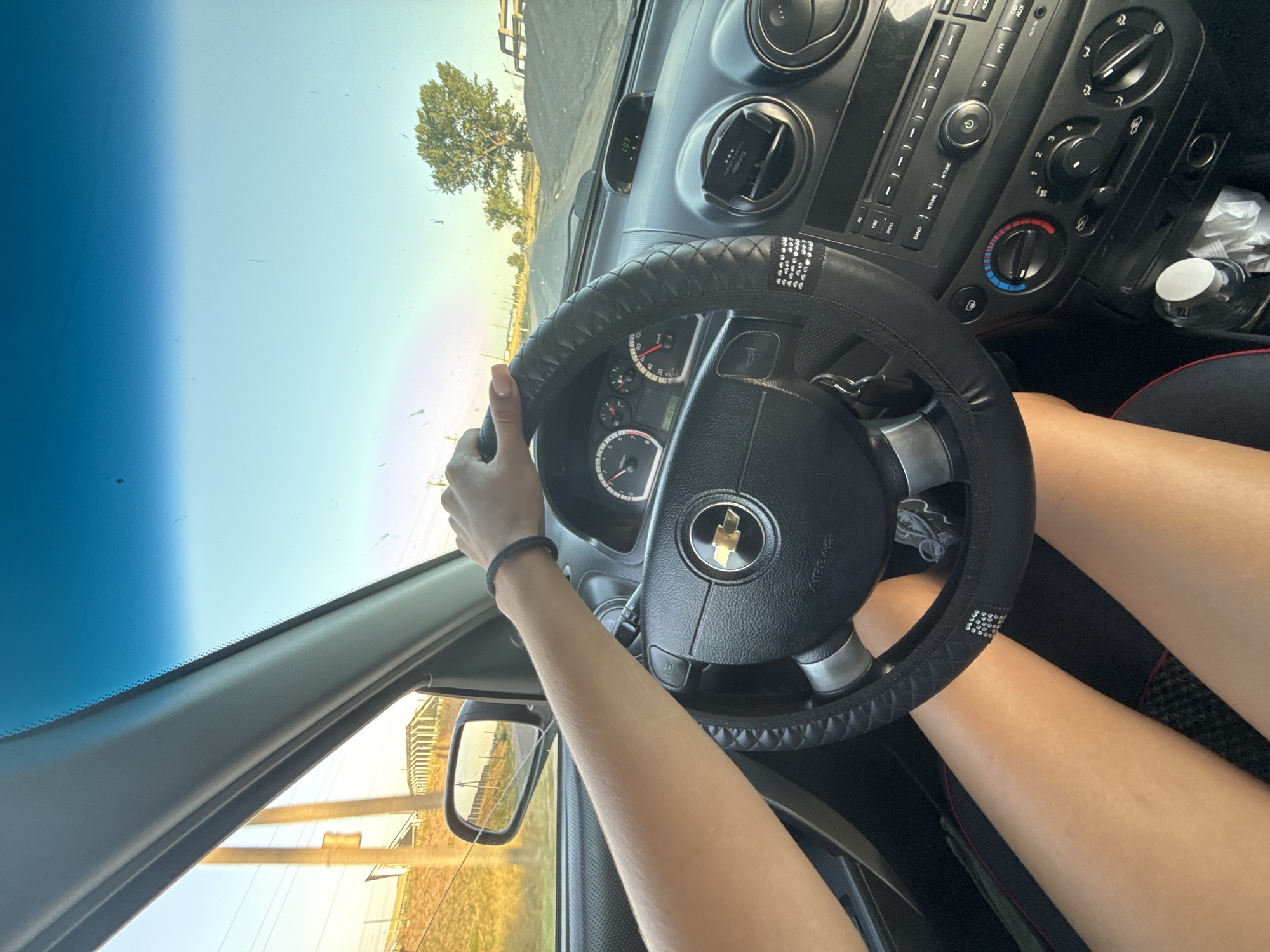 The image shows a first-person perspective from inside a car, with a person's hands on the steering wheel and their legs visible. The car appears to be in motion on a paved road. The exterior view through the windshield reveals a rural or semi-rural landscape with a clear sky. There are some utility poles and a distant tree visible. The car's dashboard is also in view, showing the speedometer and other controls. The scene suggests a daytime journey. There is no specific indication of the location within Cimișlia, Moldova, in the image itself.