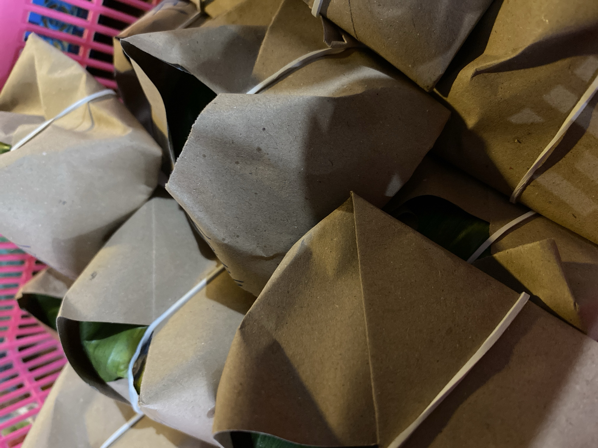 A close-up, overhead shot shows many brown paper-wrapped food packets arranged in a pink plastic basket. A white rubber band secures each packet. Some green material is visible peeking out from the top of a few packets. The food appears to be prepared for takeaway or distribution. The scene is likely in Kasihan, Indonesia, given the context.