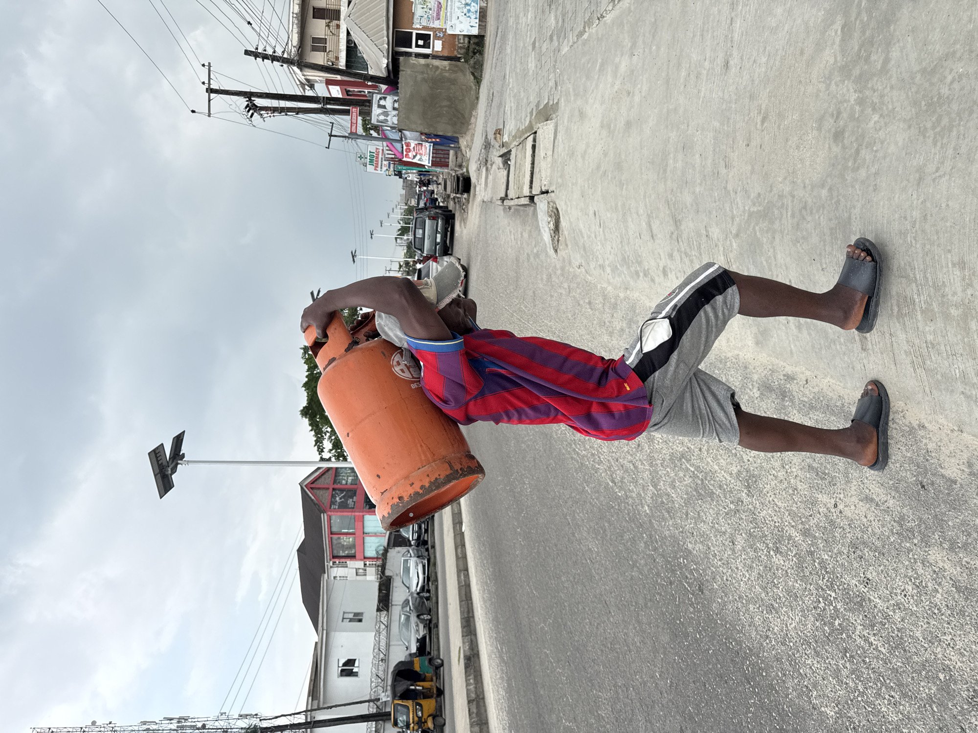 In the city of Ado, Nigeria, a man walks on the sidewalk of a street, carrying a large orange gas cylinder balanced on his right shoulder. He is wearing a striped red and purple t-shirt and grey shorts with black and white stripes. He has black skin and is wearing black sandals. The street is paved and appears to be in an urban area. There are several cars and a yellow and green motorized rickshaw on the street. Buildings with various colors and styles line the street, and utility poles with wires are visible. The sky is overcast.