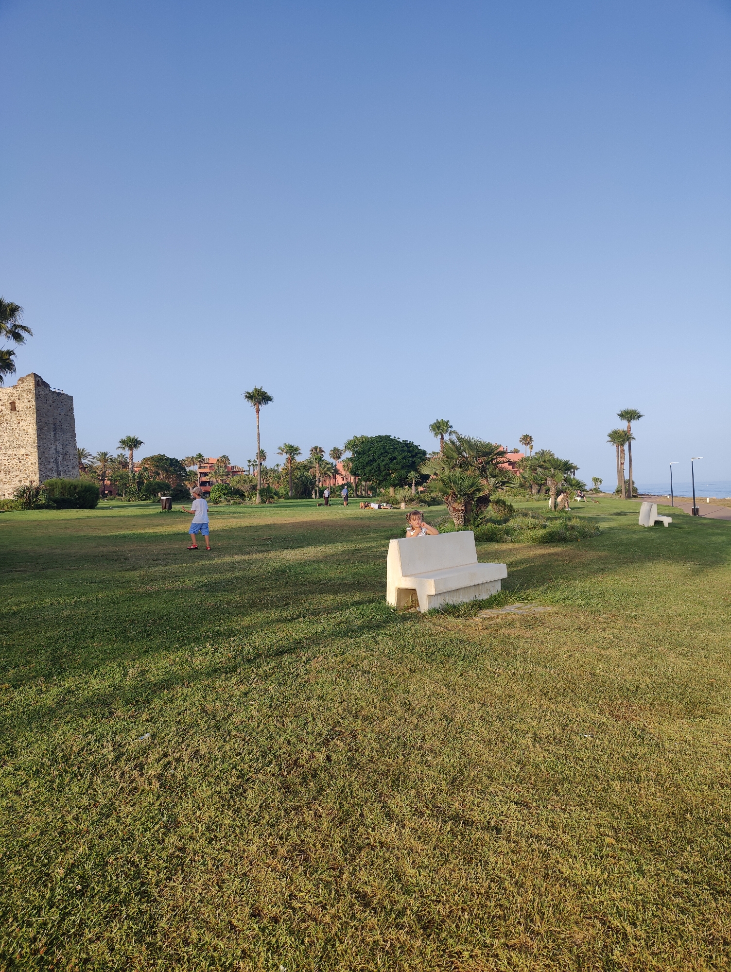 An outdoor wide shot captures a park with a clear blue sky. In the foreground, a white concrete bench sits on a green lawn. To the left of the bench, a young person wearing a white shirt and blue shorts stands on the grass. Further in the background, several palm trees are scattered throughout the park. Behind the trees, buildings with terracotta roofs are visible. To the far left of the image, a stone tower stands against the sky. In the middle ground, a young child is sitting on the white bench, looking towards the camera. In the far distance, a body of water, possibly the sea, is visible.