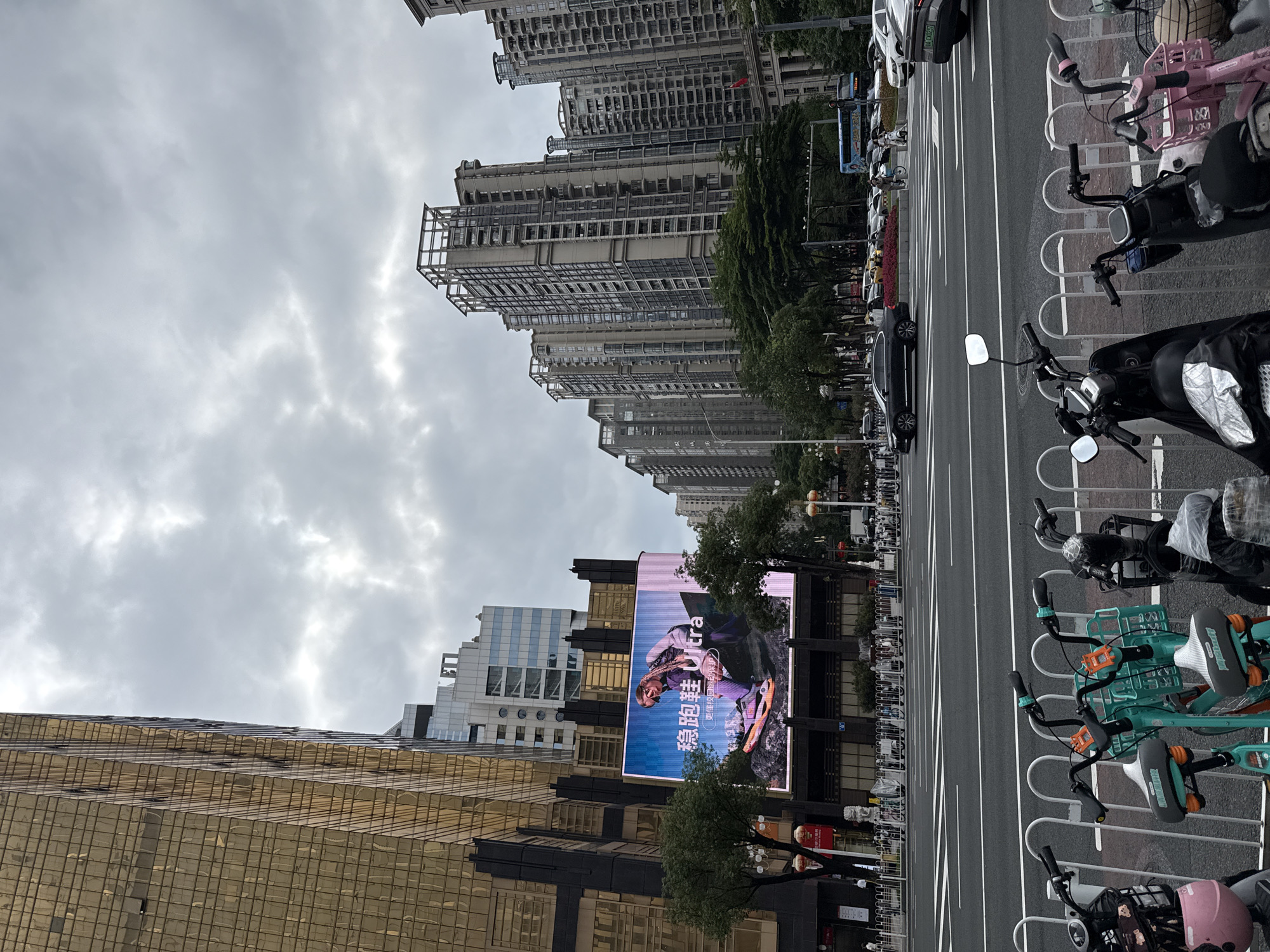 This is a medium-distance, eye-level shot of a street scene in Tianhe District, China. The foreground features a row of parked electric scooters and bicycles. Beyond them is a multi-lane road with several cars and a bus in motion. Across the road, a large digital billboard displays an advertisement for running shoes. To the left of the billboard is a tall, gold-colored building. Further in the background, numerous high-rise residential buildings dominate the skyline under a cloudy sky. Trees and streetlights are visible along the sidewalks. The overall impression is of a busy urban environment.