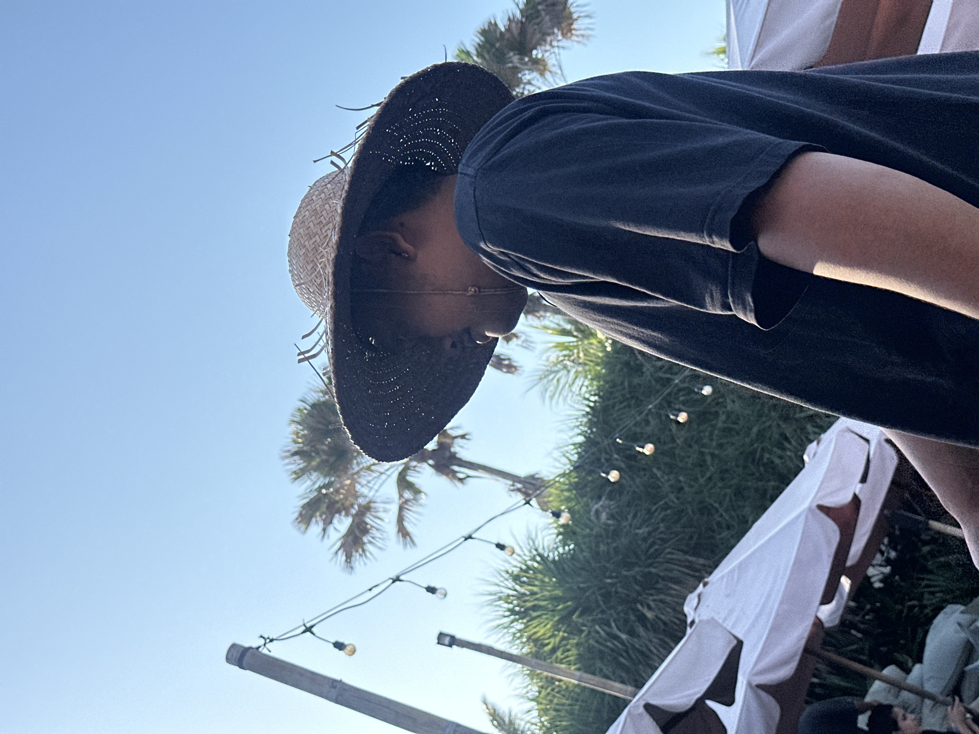 A person wearing a wide-brimmed straw hat and a dark t-shirt is shown in profile against a clear blue sky. Palm trees and lush green foliage are visible in the background. String lights with exposed bulbs are also present, adding to the outdoor ambiance. In the foreground, white umbrellas, possibly for shade, are partially visible. The scene appears to be an outdoor venue, possibly a restaurant or resort, in North Kuta, Indonesia, indicated by the tropical vegetation and clear sky.