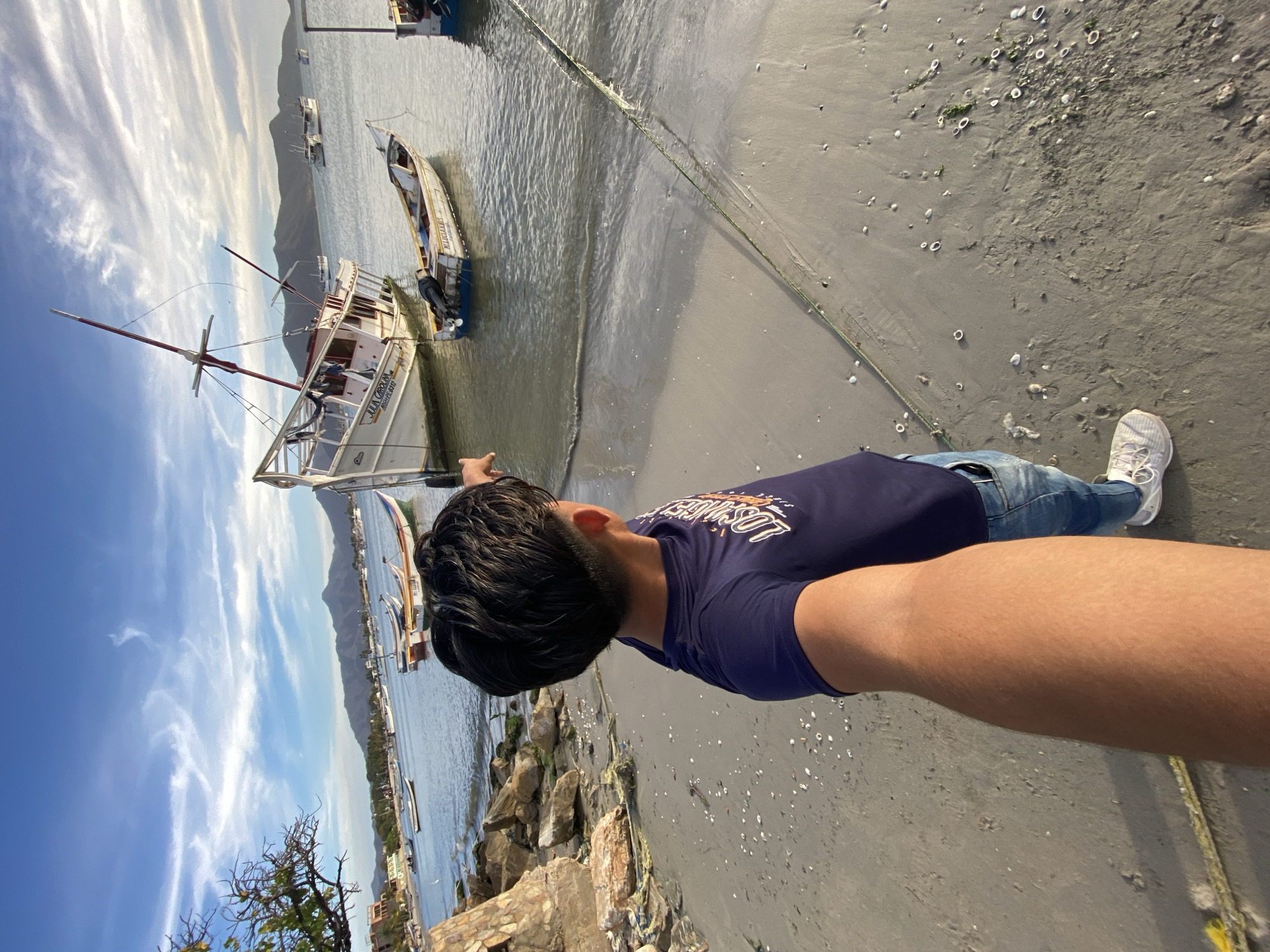 A first-person perspective captures a man on a beach in Juan Griego, Venezuela, pointing toward a boat that is beached next to the water. The man is wearing a dark blue shirt, jeans, and white sneakers. The beach is composed of gray sand, and seashells and rocks are scattered across the scene. The water is calm, and other boats are visible in the distance. The sky shows a mix of blue and white clouds. Buildings are visible in the background along the coast. The man's right arm is extended towards the camera.