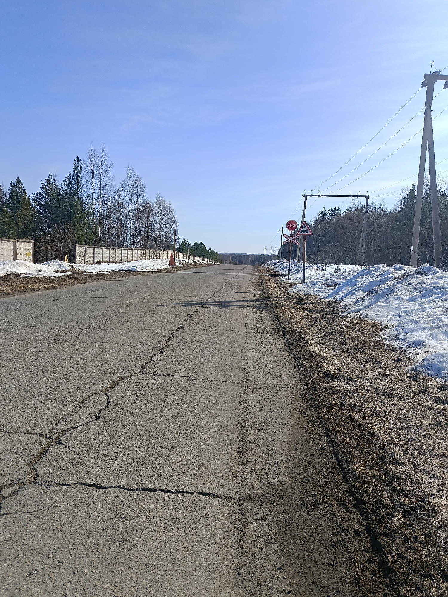 The image depicts a road in Chaikovsky, Russia. The asphalt road is cracked and shows signs of wear. On the left side of the road is a concrete fence with patches of melting snow. An orange traffic cone is visible near the fence. The right side of the road features a dirt shoulder with residual snow cover. A utility pole stands on the right side of the road along with a railroad crossing sign and a stop sign. The background shows a forest and a clear blue sky.