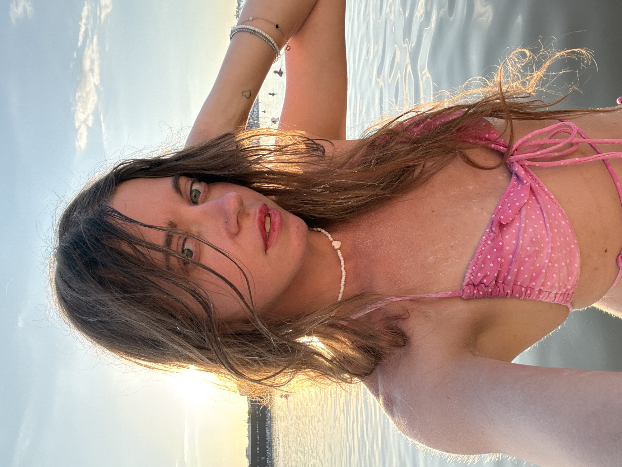 A young woman with long, wavy brown hair is taking a selfie in the water at sunset in Sol-Iletsk, Russia. She is wearing a pink polka dot bikini and a white beaded necklace. Her right arm is raised, with her hand behind her head. She has a small heart tattoo on her left forearm and is wearing bracelets on her right wrist. The sun is low in the sky, casting a warm glow on the water and the woman's face. In the background, there are silhouetted figures of people swimming and a distant cityscape. The water appears calm with gentle ripples.