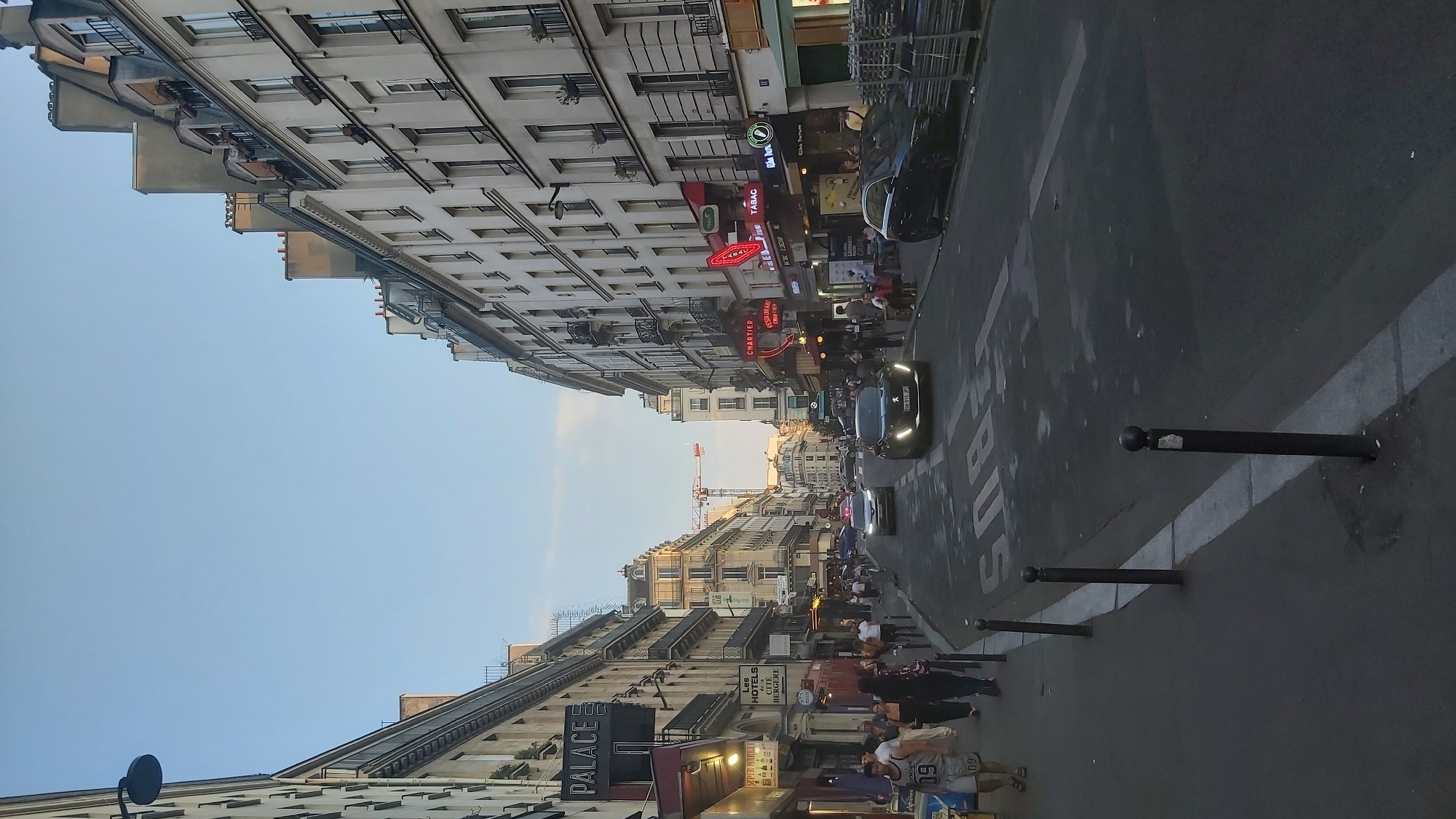 This is a street view in Paris, France, likely taken in the late afternoon or early evening. The street is lined with multi-story buildings typical of Parisian architecture, featuring cream-colored facades, numerous windows, balconies, and pitched roofs with dormer windows. The street is paved and has white lane markings, including a prominent "BUS" marking. Several cars are visible on the street, including a dark sedan and a silver sedan. Pedestrians are walking on the sidewalk, which is separated from the street by bollards. Some of the buildings have illuminated signs for businesses such as "PALACE," "SUPER MARKET," "HOTELS," "CHARTIER," "TABAC," and a restaurant. A construction crane is visible in the distance, suggesting ongoing development or renovation in the area. The sky is clear and blue, with a hint of a gradient towards the horizon.