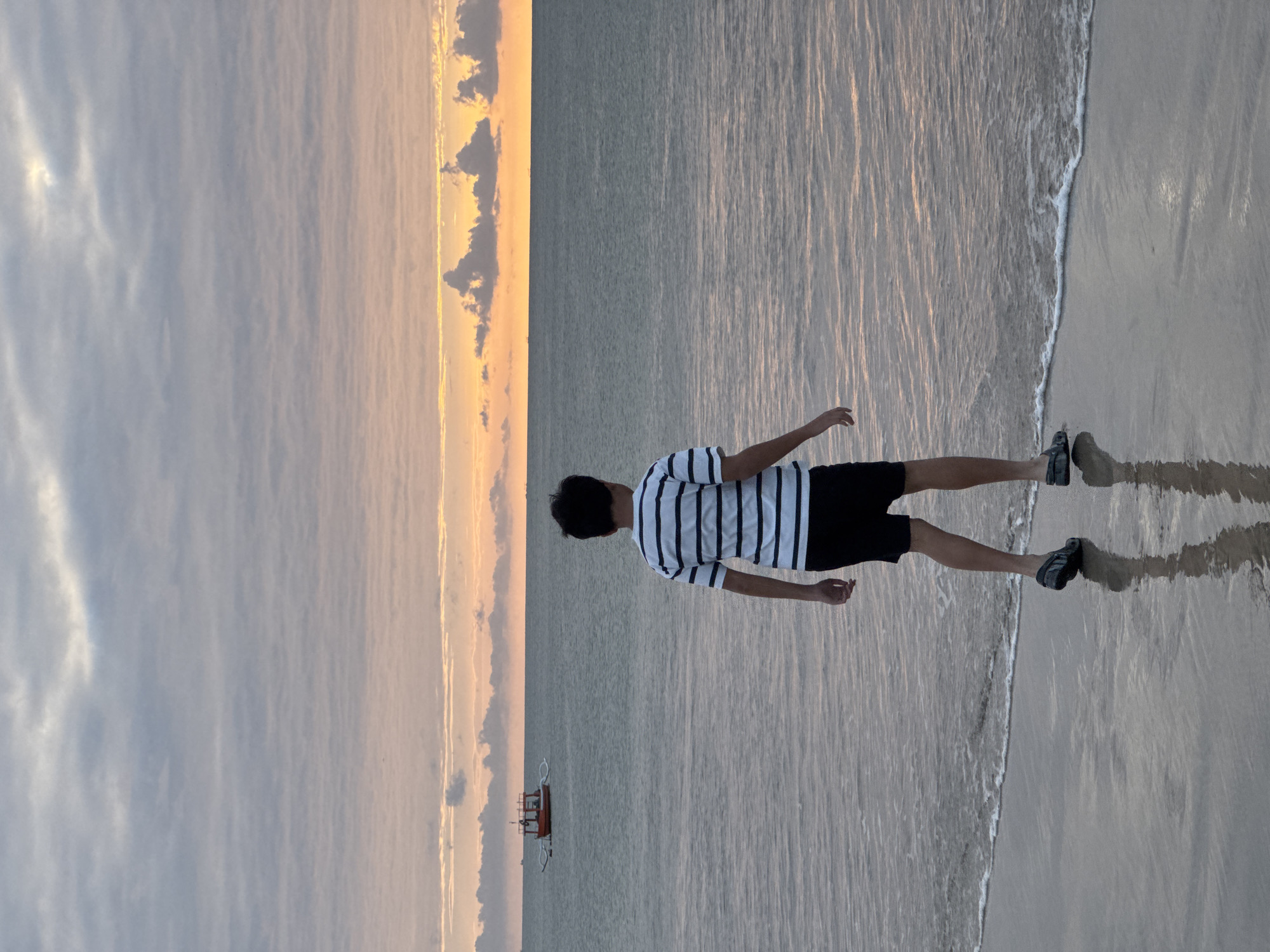 A young person stands on a sandy beach, facing the water. They are wearing a black and white striped t-shirt and black shorts, with dark shoes. The person's reflection is visible on the wet sand, distorted by the shallow water lapping at their feet. The sun is low in the sky, casting a warm glow on the horizon, where clouds are tinged with orange and yellow. The sea is calm, with gentle waves breaking on the shore. In the distance, a small boat with a red hull is visible on the water. The overall atmosphere is serene and peaceful, suggesting a moment of quiet contemplation at sunset. The location appears to be a tropical beach, possibly in Kuta Selatan, Indonesia, given the warm hues of the sky and the presence of the boat. No visible text is present in the image.