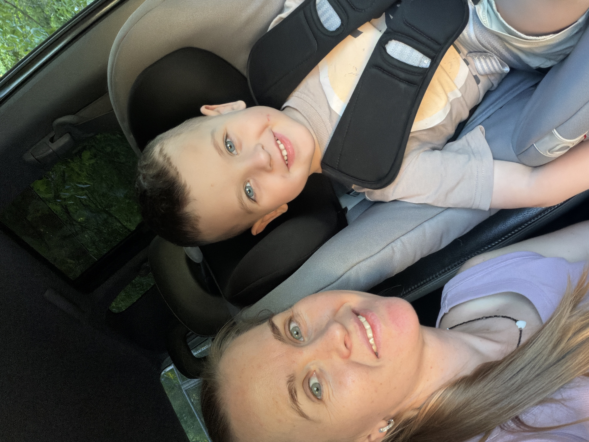 A medium shot captures a woman and a young boy in a car. The woman, with blonde hair pulled back in a ponytail, faces the camera with a smile, her light blue eyes visible. She wears a light purple tank top and a necklace. To her right, the boy, with short brown hair and bright blue eyes, is seated in a car seat, smiling directly at the camera. He is wearing a light brown t-shirt and the car seat's black harness system is across his chest. The background shows the interior of a car and blurred greenery through the windows, suggesting an outdoor environment. The scene appears to be a selfie taken from the driver's perspective. The location of Khabarovsk, Russia is not discernible from the image.