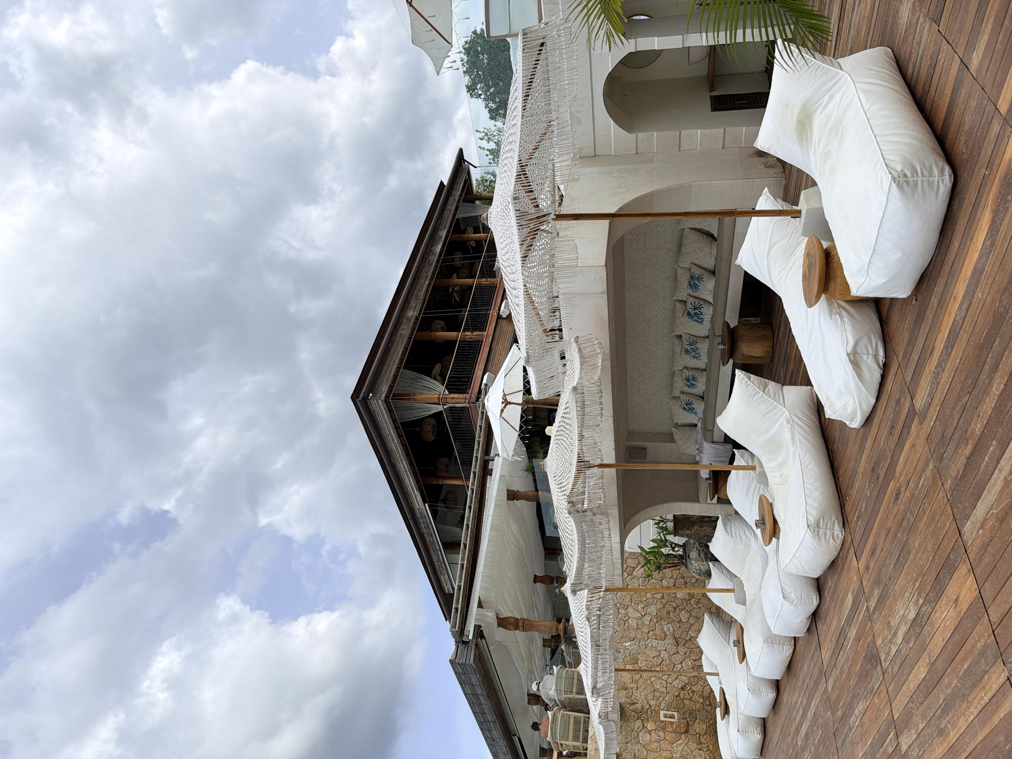 This image depicts an outdoor seating area at a resort or restaurant in Koh Samui District, Thailand. The foreground features a wooden deck with several white beanbag-style lounge chairs arranged in a row, each with a small wooden side table. Behind the lounge chairs, a white stone structure is visible, topped with a row of white umbrellas with macrame fringe. Further back, a multi-story building with a dark wooden exterior and balconies is partially visible, with white curtains hanging from the windows. The background shows a cloudy sky. The scene appears to be tranquil and set up for relaxation or dining.