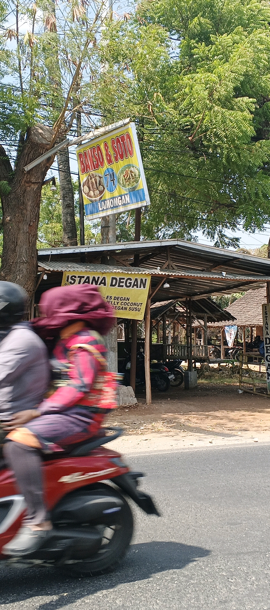 The image captures a street scene in Kandangsemangkon, Indonesia, dominated by a roadside eatery. A red motorcycle with two blurred occupants, one wearing a maroon head covering and a patterned top, is in motion in the foreground. Behind the motorcycle, a yellow banner reads "ISTANA DEGAN" and lists menu items like "ES DEGAN," "JELLY COCONUT," and "DEGAN SUSU." Above this, a blue and yellow sign advertises "BAKSO & SOTO LAMONGAN." The eatery itself features a rustic, open-air structure with a corrugated metal roof supported by wooden beams. Several motorcycles are parked in the background under the shelter. The environment appears to be a rural or suburban setting with lush green trees and a bright, sunny sky.