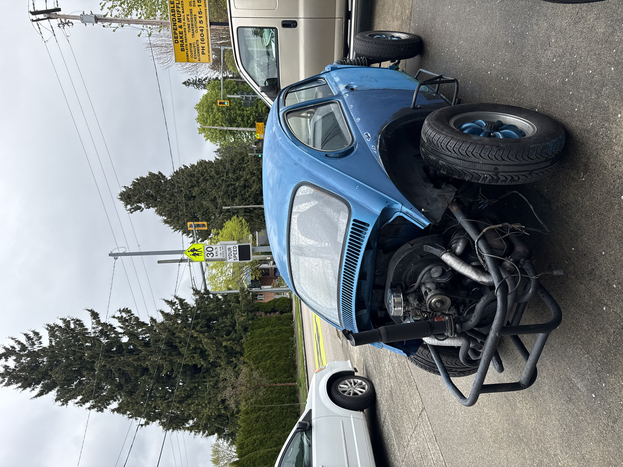 The image, taken in New Westminster, Canada, captures a street scene on an overcast day, with visible water droplets suggesting recent rain. The central subject is a distinctive light blue, heavily modified car resembling a Volkswagen Beetle, possibly a dune buggy conversion, parked on the asphalt. Its front end is open, revealing an engine and a custom black tubular frame/bumper. The car features blue-painted wheel hubs and appears to be a work-in-progress or custom build.
Partially visible on either side are two other vehicles: a white minivan to the left and a gold/tan van to the right. The background includes power poles with numerous utility wires, lush green trees, and bushes lining the street. Traffic lights are visible, with one showing green and another yellow.
Notable road signage includes a yellow diamond-shaped sign on a pole, indicating a "30 km/h" speed limit for a "SCHOOL" zone between "7 AM-10 PM," with a "YOUR SPEED" sign below it. Further back, a large yellow sign is attached to a utility pole, advertising "DEPENDABLE BRAKE & MUFFLER." This sign details services such as "COMPLETE REPAIRS TO ALL MAKES," "TUNE UP'S (CUSTOM EXHAUST)," "TRANSMISSIONS OIL," "ALIGNMENTS CLUTCHES," and provides a phone number: "PH (604) 515-15," concluding with "YOUR NEIGHBOURHOOD AUTOMOTIVE SERV." The scene depicts an urban or suburban setting.