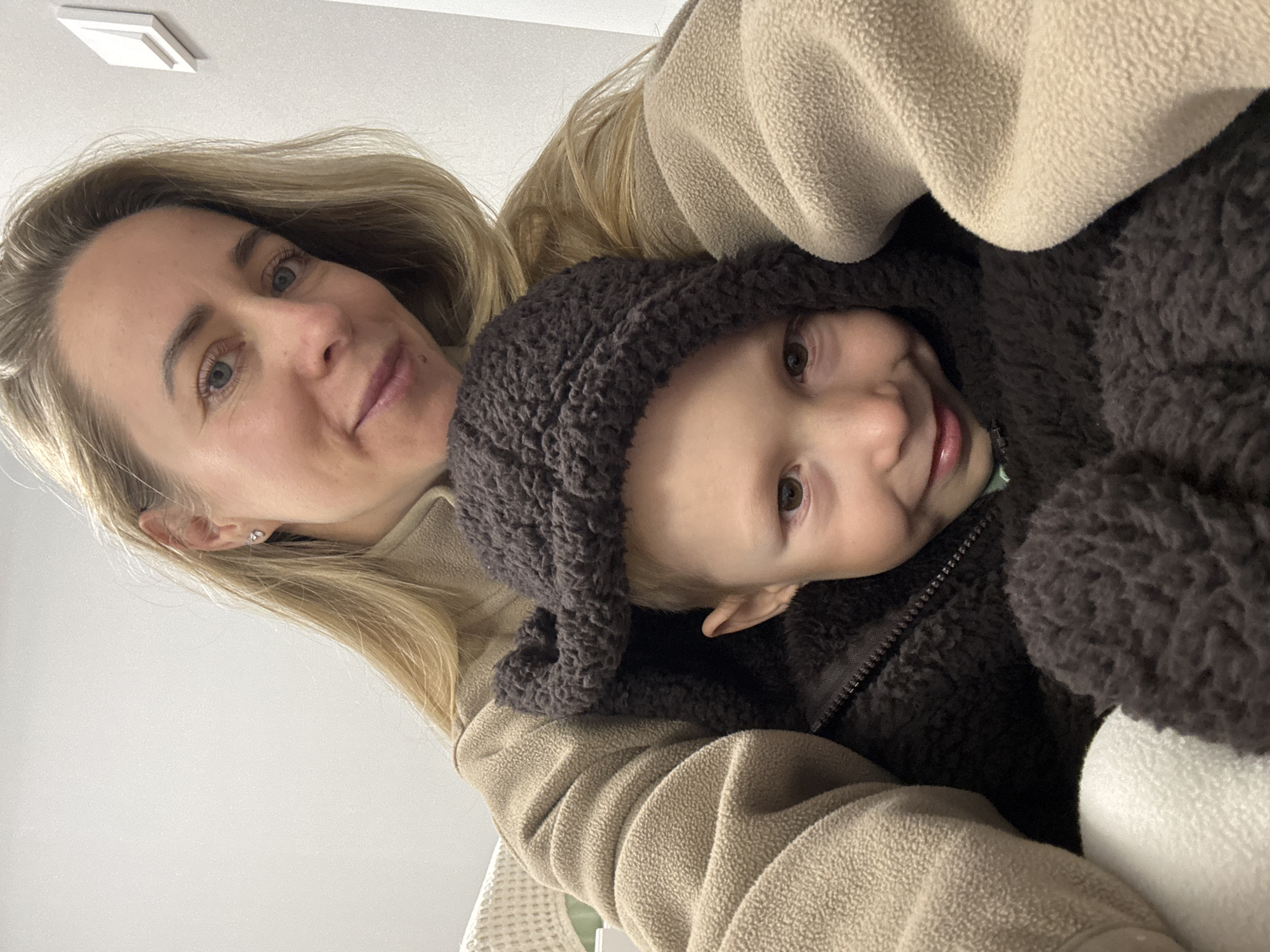 A selfie taken from a low angle shows a woman and a child. The woman, on the right, has blonde hair and is wearing a light brown fleece jacket. She is looking towards the camera with a slight smile. The child, on the left, is wearing a dark brown furry hooded outfit and is peeking out from under the woman's chin. The child also has a smile on their face. The background is a plain, light-colored wall with a white geometric object mounted on it, possibly a light switch or decorative item. The overall setting appears to be indoors, in what could be a waiting room or home environment. No specific location in Chaikovsky, Russia can be determined from the visual information provided in the image.