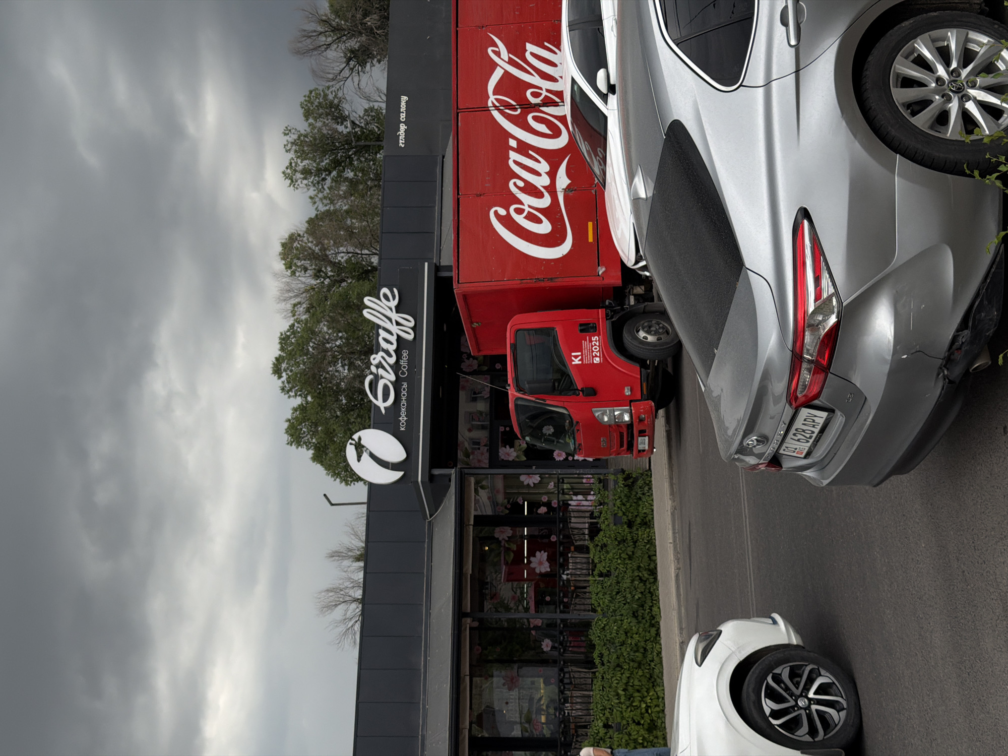 An overcast, cloudy sky looms over a street scene. On the right, a red Coca-Cola truck is parked next to a white car, with a silver car in the foreground. To the left, a red truck is parked in front of a building with a "Giraffe Coffee" sign. The building has large windows adorned with floral patterns and a green hedge runs along the sidewalk. A person stands on the left side of the frame, facing away from the camera.