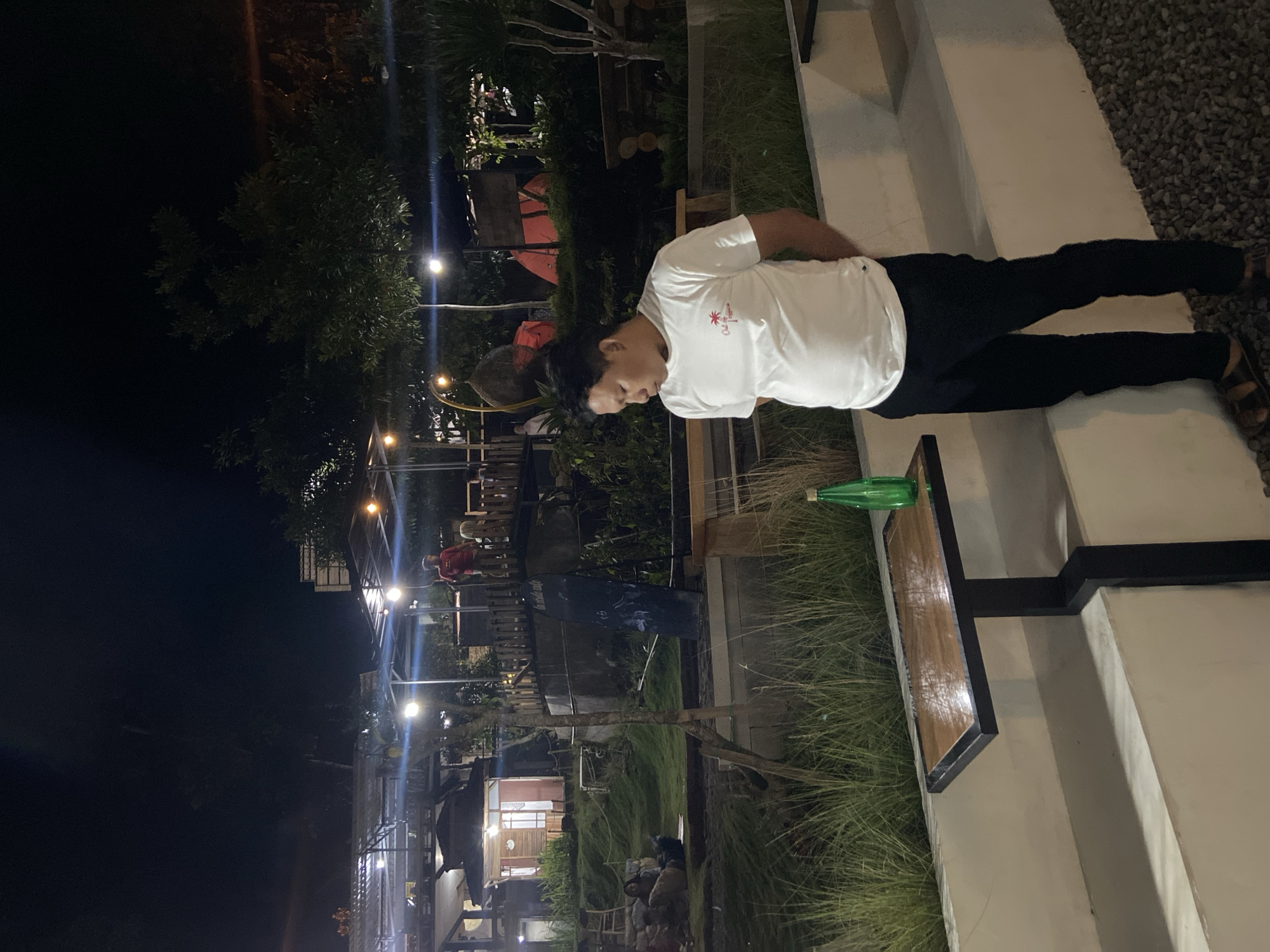 The image depicts an outdoor scene at night, likely a cafe or restaurant with a rustic ambiance, possibly located in the city of Kesiman, Indonesia. In the foreground, a man wearing a white t-shirt and black pants stands facing away from the camera, his hands clasped behind his back. He is positioned next to a small, dark-legged table with a wooden top, on which a green bottle is placed.
The background reveals a multi-level structure with open-air seating areas, illuminated by warm lights. Some buildings with windows are visible, suggesting habitation or service areas. In the mid-ground, lush green grass slopes downward, with people seated or lounging on it. Further back, trees and a red tent are discernible, contributing to a natural and perhaps camping-like setting. The overall atmosphere appears relaxed and casual, with the night lights creating a warm and inviting glow. The ground in the immediate foreground is covered with small pebbles.