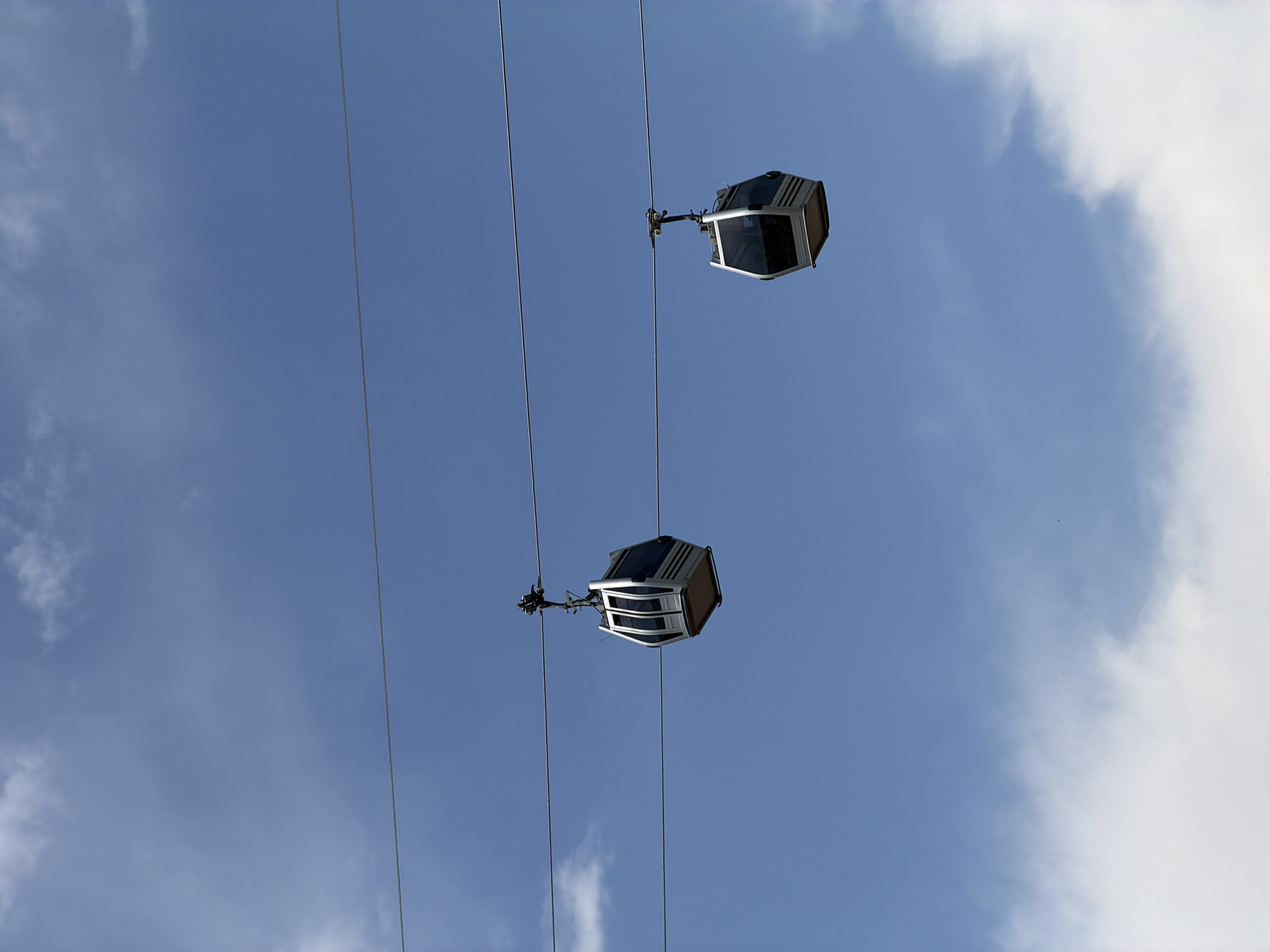 This image depicts two cable cars suspended from wires against a blue sky with scattered white clouds. The cable cars are modern, hexagonal in shape, with silver exteriors and tinted windows. They are positioned at different points along the wires, suggesting movement or parallel lines of travel. The scene is likely an outdoor urban environment, possibly a tourist attraction or a mode of public transportation. The presence of cable cars in Tbilisi, Georgia, strongly suggests a location such as the Narikala Fortress or the Rike Park area, which are known for their aerial tramway systems offering scenic views of the city.